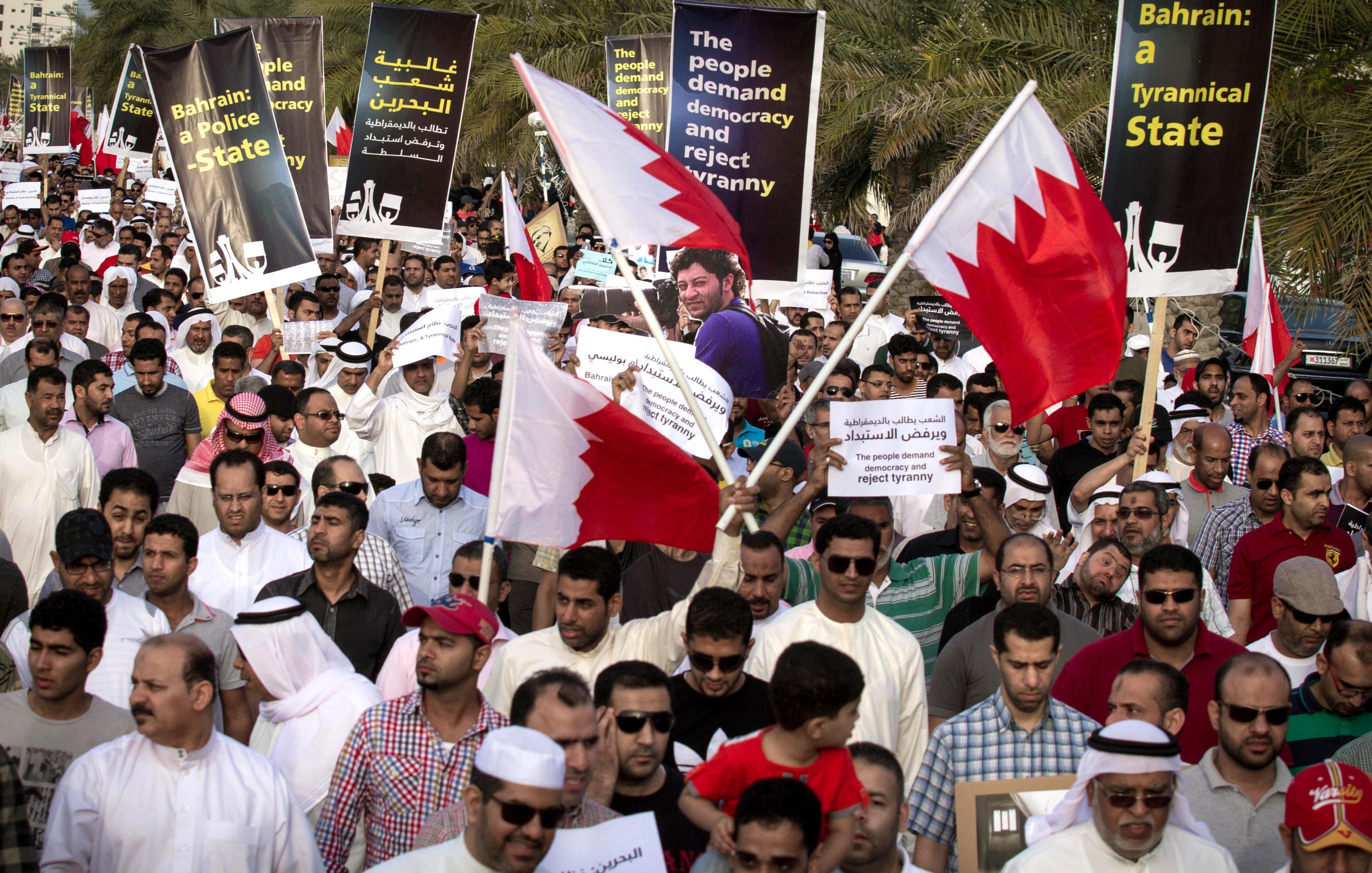 الصورة تظهر تجمعًا حاشدًا لمتظاهرين في البحرين يحملون لافتات وأعلام البلاد. يتواجد الأشخاص في مكان مفتوح، ويسود الجو شعور بالتضامن والمطالبة بالتغيير. اللافتات تتضمن مطالب للديمقراطية ورفض السياسات القمعية. يرتدي المتظاهرون ملابس تقليدية وطنية، ويبدو أن الحشد متنوع من حيث الأعمار.