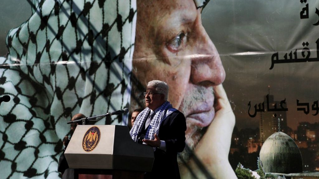 L'image représente un homme en train de parler lors d'un discours. Il se tient devant un grand poster qui affiche le portrait d'un homme en keffieh, regardant pensivement au loin. Le décor suggère qu'il s'agit d'un événement commémoratif, probablement lié à des thèmes politiques ou historiques. Le podium sur lequel l'homme s'exprime est orné d'un emblème officiel, tandis que le fond montre une ville, ajoutant un aspect symbolique à la scène.