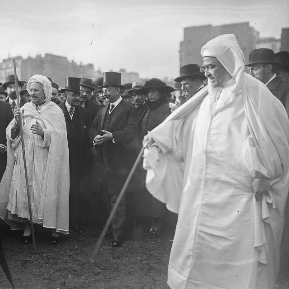 L'image montre un groupe de personnes dans un environnement urbain. Au centre, plusieurs individus portent des vêtements traditionnels, tels que des robes blanches et des turbans. D'autres, habillés de manière formelle avec des chapeaux haut de forme et des costumes, se tiennent autour d'eux, suggérant une rencontre entre différentes cultures. L'arrière-plan présente des bâtiments typiques de l'époque. L'atmosphère semble être celle d'un événement officiel ou d'une cérémonie.