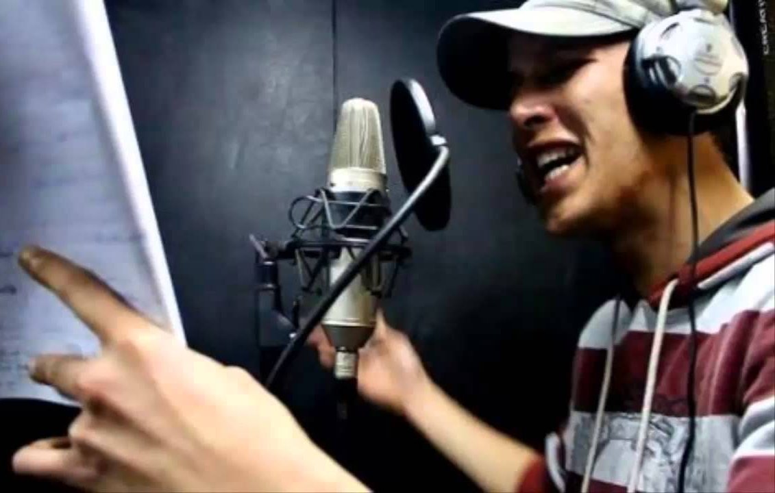 L'image montre un homme en train de chanter ou de rapper dans un studio d'enregistrement. Il porte un chapeau et des écouteurs. Devant lui, un microphone est connecté et il tient des feuilles de papier, probablement des paroles ou un texte. L'expression de son visage indique qu'il est très impliqué dans sa performance. L'arrière-plan semble sombre, ce qui est typique des environnements d'enregistrement.
