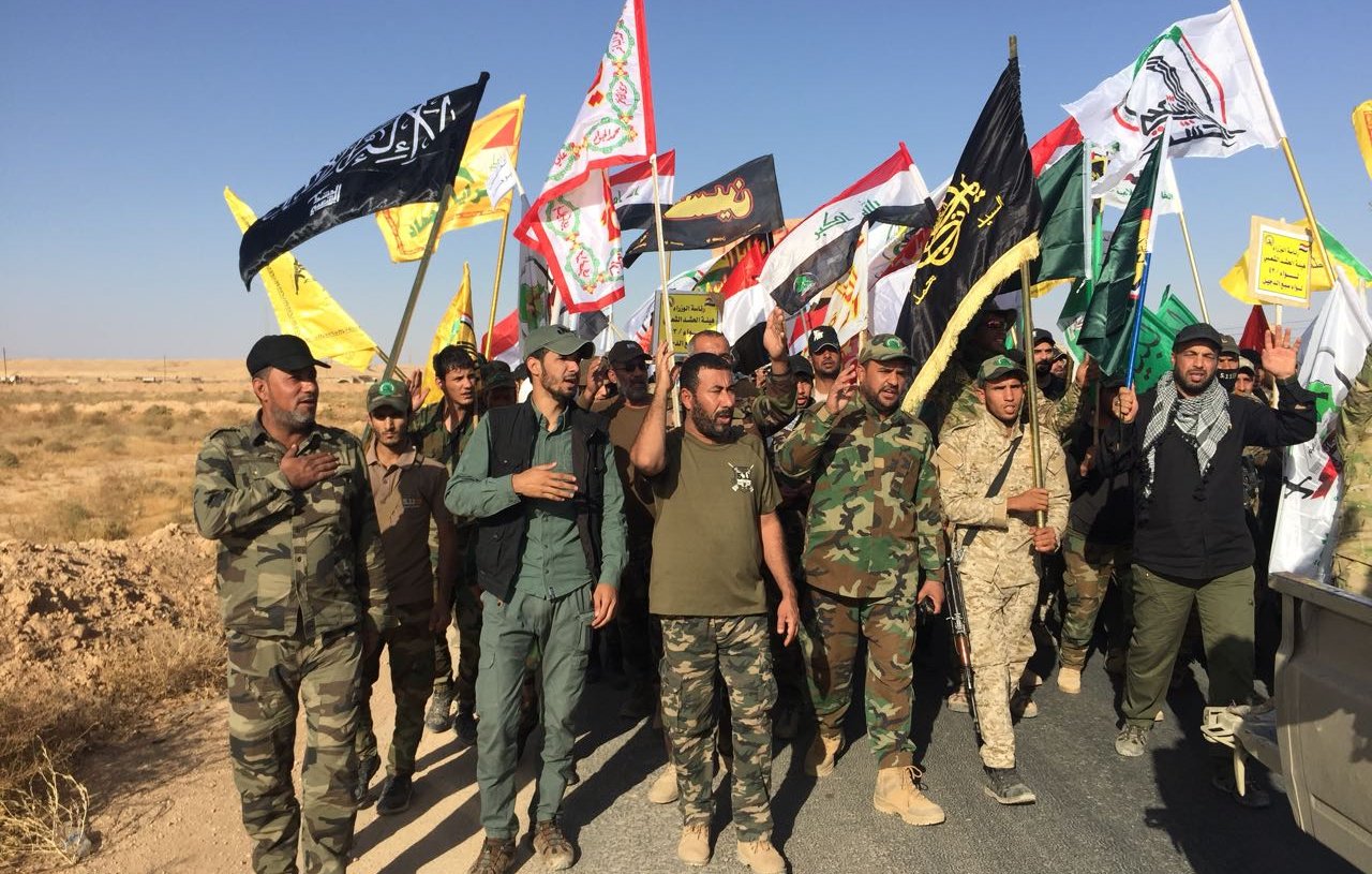 The image depicts a group of men participating in a march or rally, holding various flags. The men are dressed in military-style clothing and appear to be organized, with some wearing uniforms and others in casual attire. The flags they carry include a mix of colors and designs, suggesting they represent different factions or groups. The scene is set in an open area, likely with dry or arid terrain in the background, and the sky is clear. The atmosphere seems to indicate a demonstration or a show of solidarity among the participants.