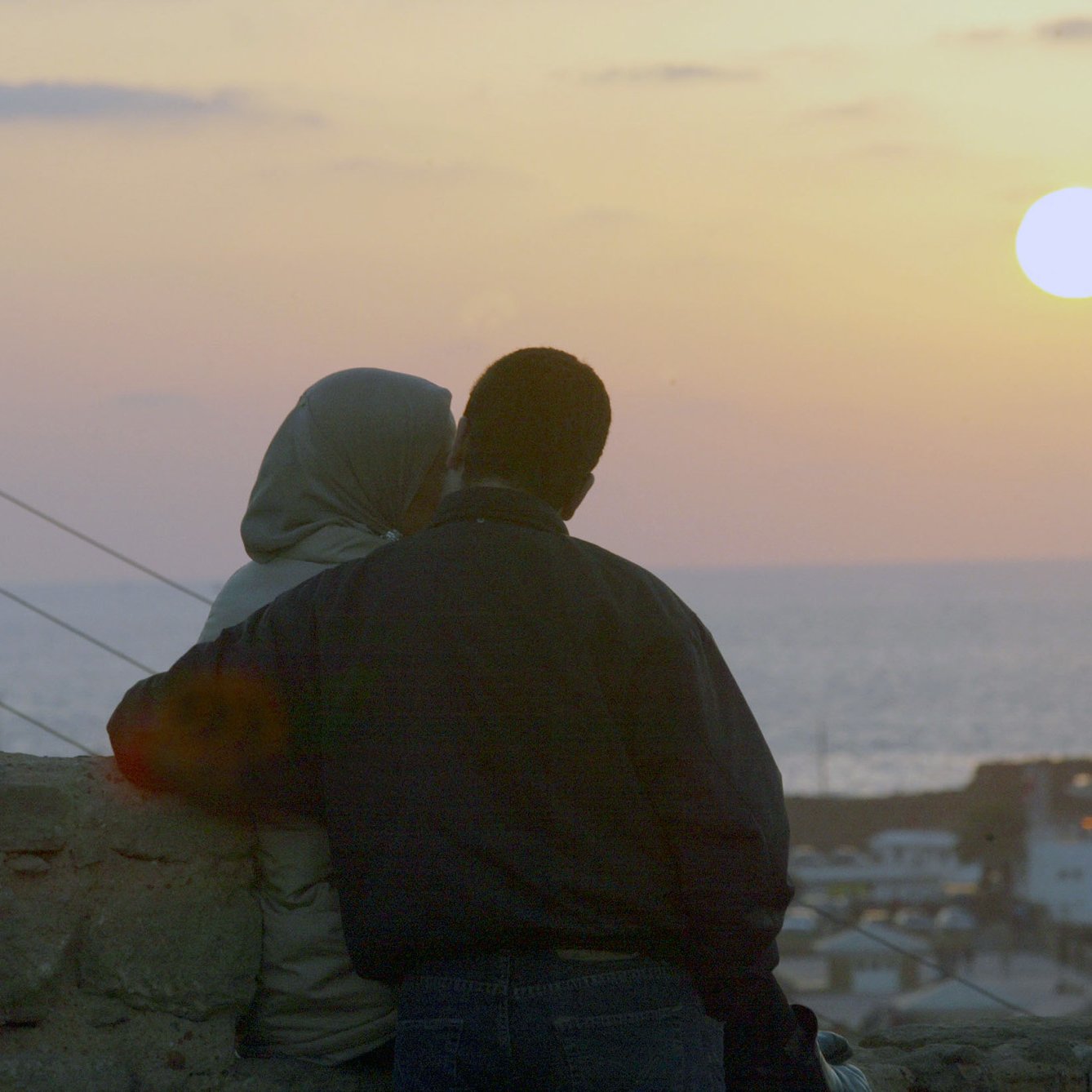 L'image montre un couple assis côte à côte, observant le coucher de soleil. L'un des membres du couple porte un hijab, tandis que l'autre est vêtu d'une veste sombre. Ils semblent se rapprocher l'un de l'autre, créant une atmosphère intime. En arrière-plan, on peut voir la mer, et le ciel est teinté de couleurs douces, avec le soleil se couchant à l'horizon. Les bâtiments sont légèrement visibles en bas, ajoutant à la scène romantique.