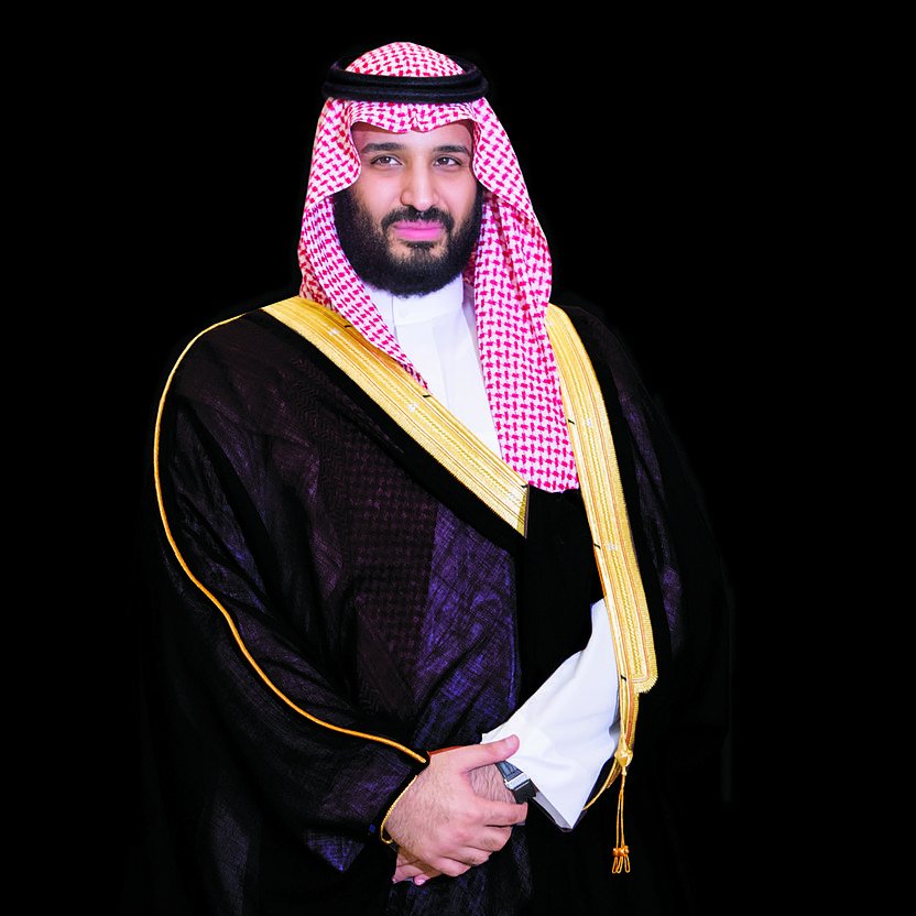 L'image montre un homme vêtu d'une tenue traditionnelle saoudienne, composée d'une darra'a noire avec des ornements dorés et d'un keffieh rouge et blanc, maintenu par une agrafe noire. Il affiche une expression sérieuse et confiante, se tenant droit avec les bras légèrement croisés devant lui. Le fond est sombre, ce qui met en valeur sa tenue et son apparence.