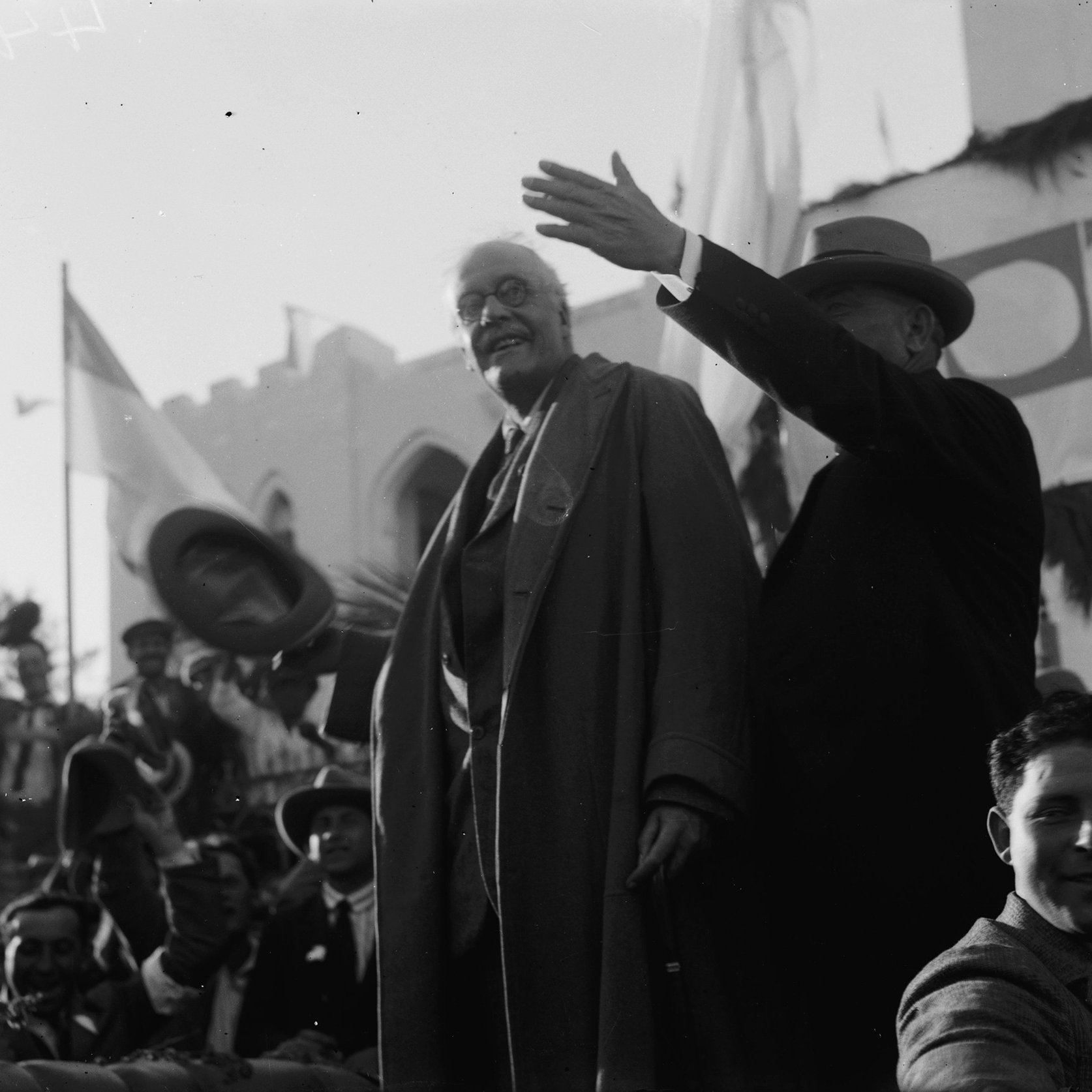L'image montre des hommes en costume debout sur une estrade, saluant une foule rassemblée. L'un des hommes, portant un manteau, semble s'adresser au public, tandis que l'autre, en chapeau, lève la main. En arrière-plan, on aperçoit des drapeaux et des décorations, indiquant un événement officiel ou une célébration. La foule est composée de personnes applaudissant et prenant part à l'événement. L'atmosphère semble festive et solennelle.