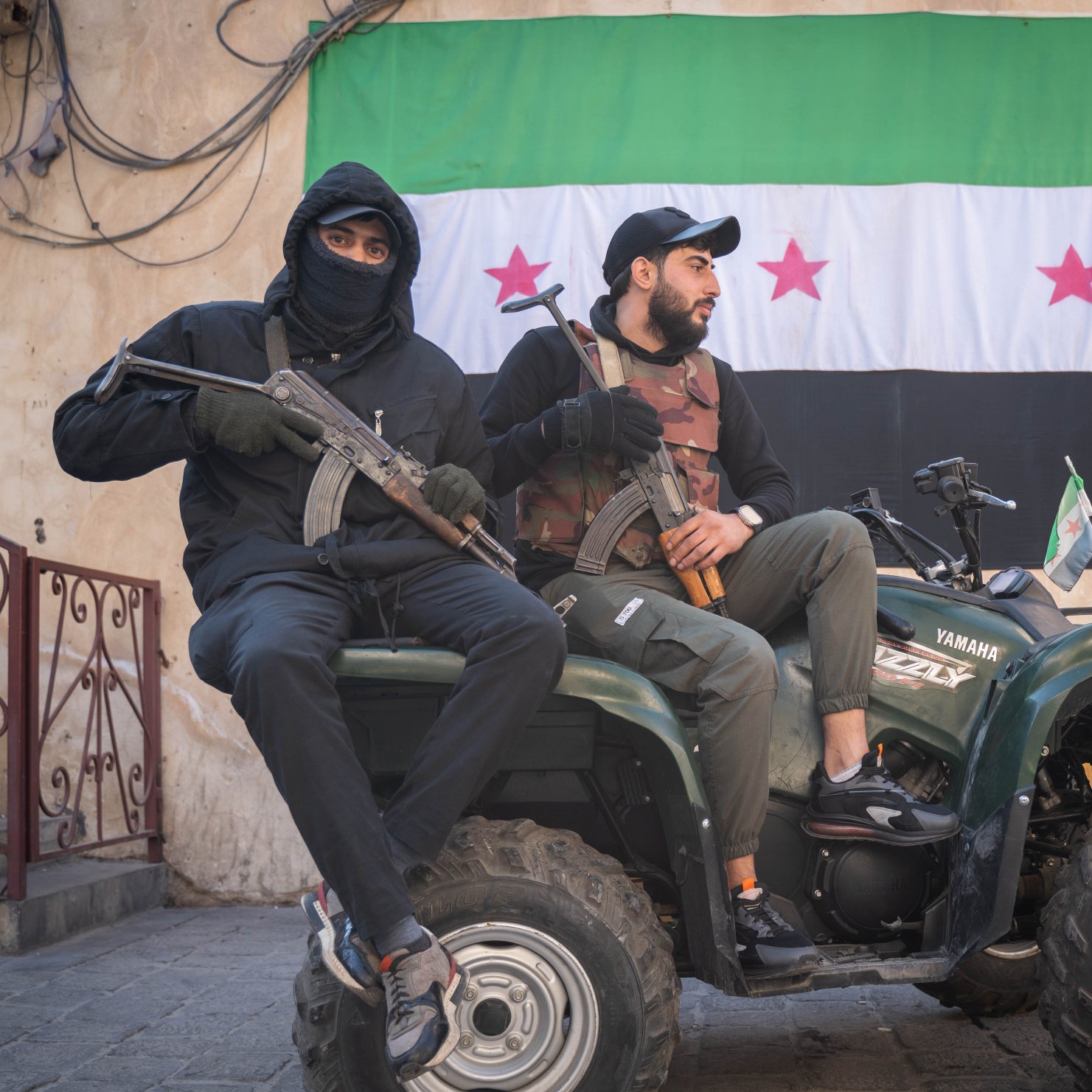 Deux hommes assis sur un quad, l'un tenant une arme, l'autre un drapeau. En arrière-plan, un mur avec un grand drapeau portant des étoiles rouges sur fond vert et noir. L'ambiance semble militaire et tendue.