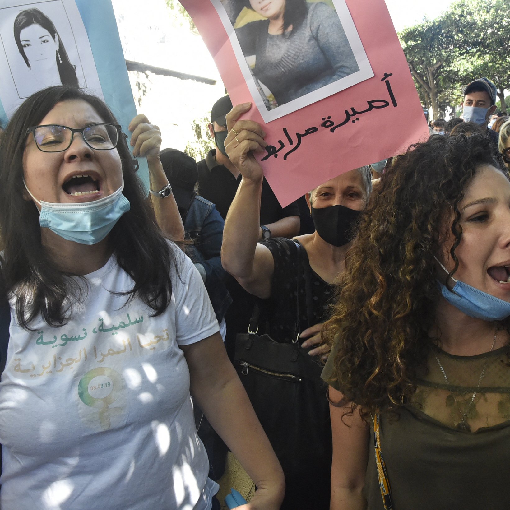 The image depicts a group of women participating in a protest. They are holding up posters, some of which feature photographs of individuals. The women appear to be chanting or shouting, expressing their emotions passionately. Many of them are wearing face masks, likely due to health precautions. The background suggests an outdoor setting with trees, indicating that the demonstration is taking place in a public space. The atmosphere seems charged with activism and determination.