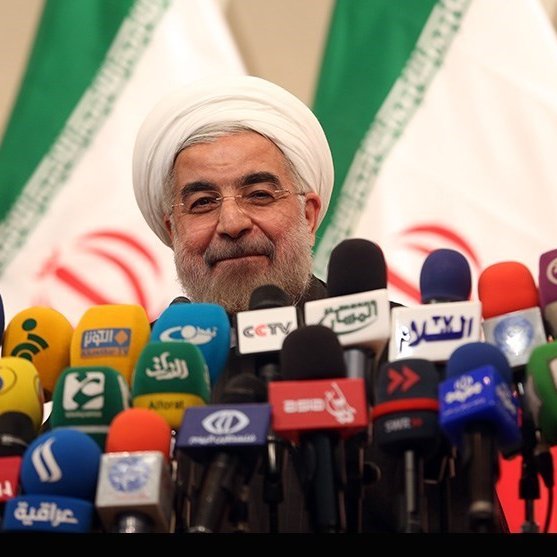 L'image montre un homme souriant, vêtu d'une tenue formelle et portant un turban blanc. Il se tient devant une multitude de microphones de différentes chaînes de télévision et médias. En arrière-plan, il y a des drapeaux avec des motifs et des couleurs qui semblent représenter un pays. L'ambiance semble être celle d'une conférence de presse ou d'un événement médiatique.