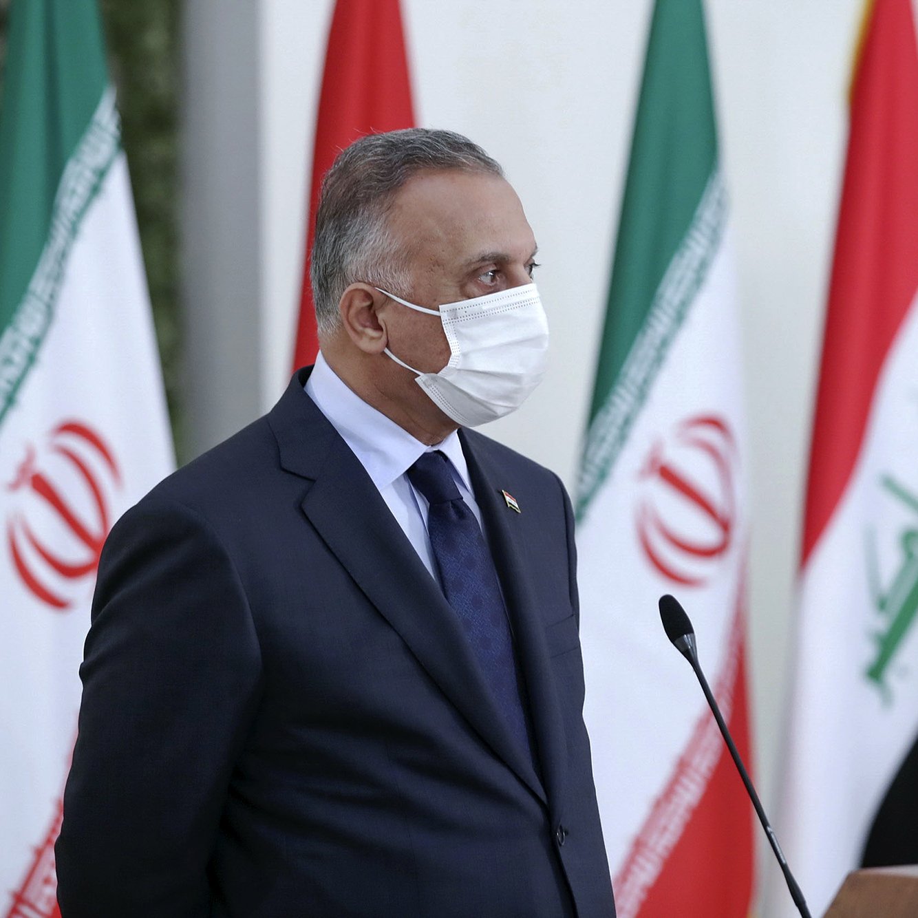 The image depicts a person wearing a mask, standing in profile. He is dressed in a dark suit with a tie and appears to be engaged in an official event. In the background, multiple flags are visible, including those of Iran and Iraq, suggesting a diplomatic or governmental setting. The person appears serious and focused, possibly addressing an audience or participating in a press conference.