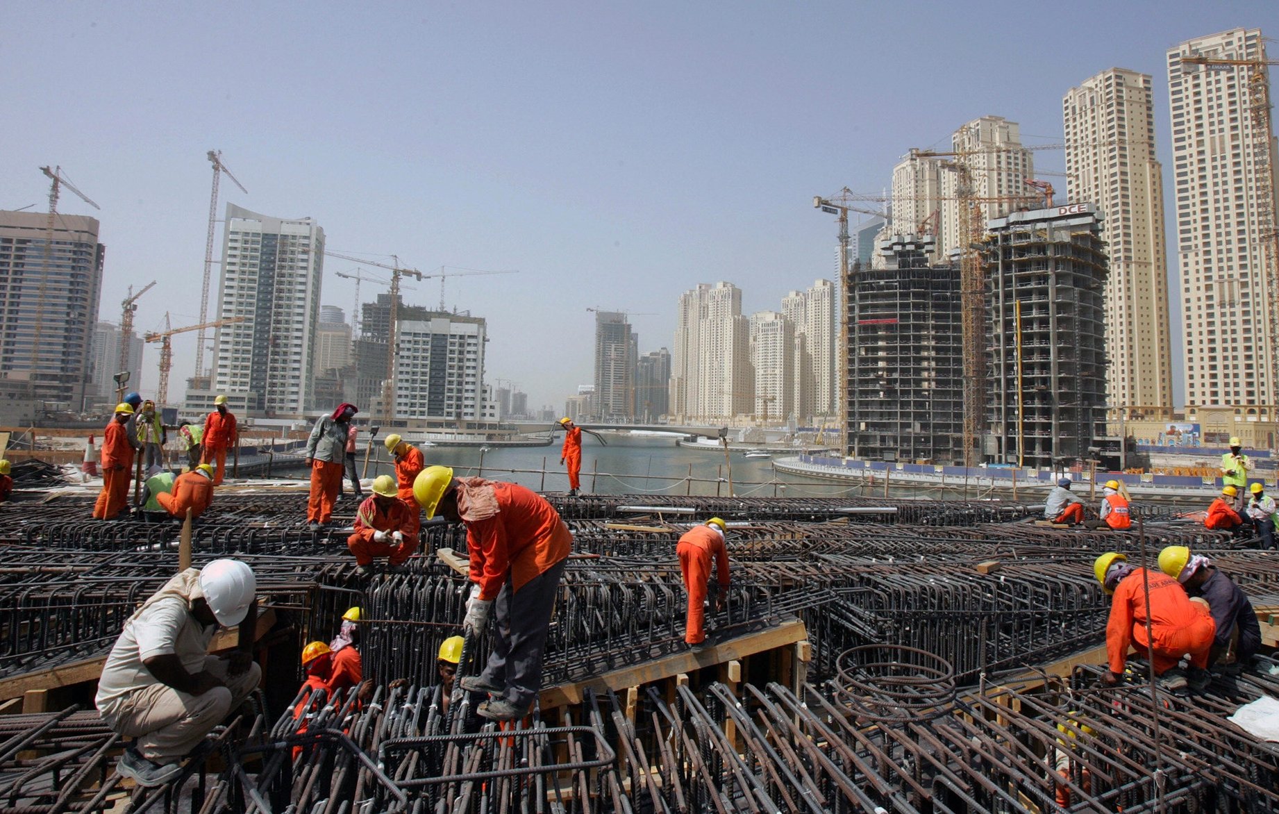 L'image montre un chantier de construction animé, où de nombreux ouvriers en uniformes orange et casques de sécurité s'affairent autour de structures en acier. En arrière-plan, on peut voir des immeubles modernes et des grues, illustrant une zone urbaine en développement. Le ciel est clair, et l'ensemble dégage une atmosphère dynamique de construction.