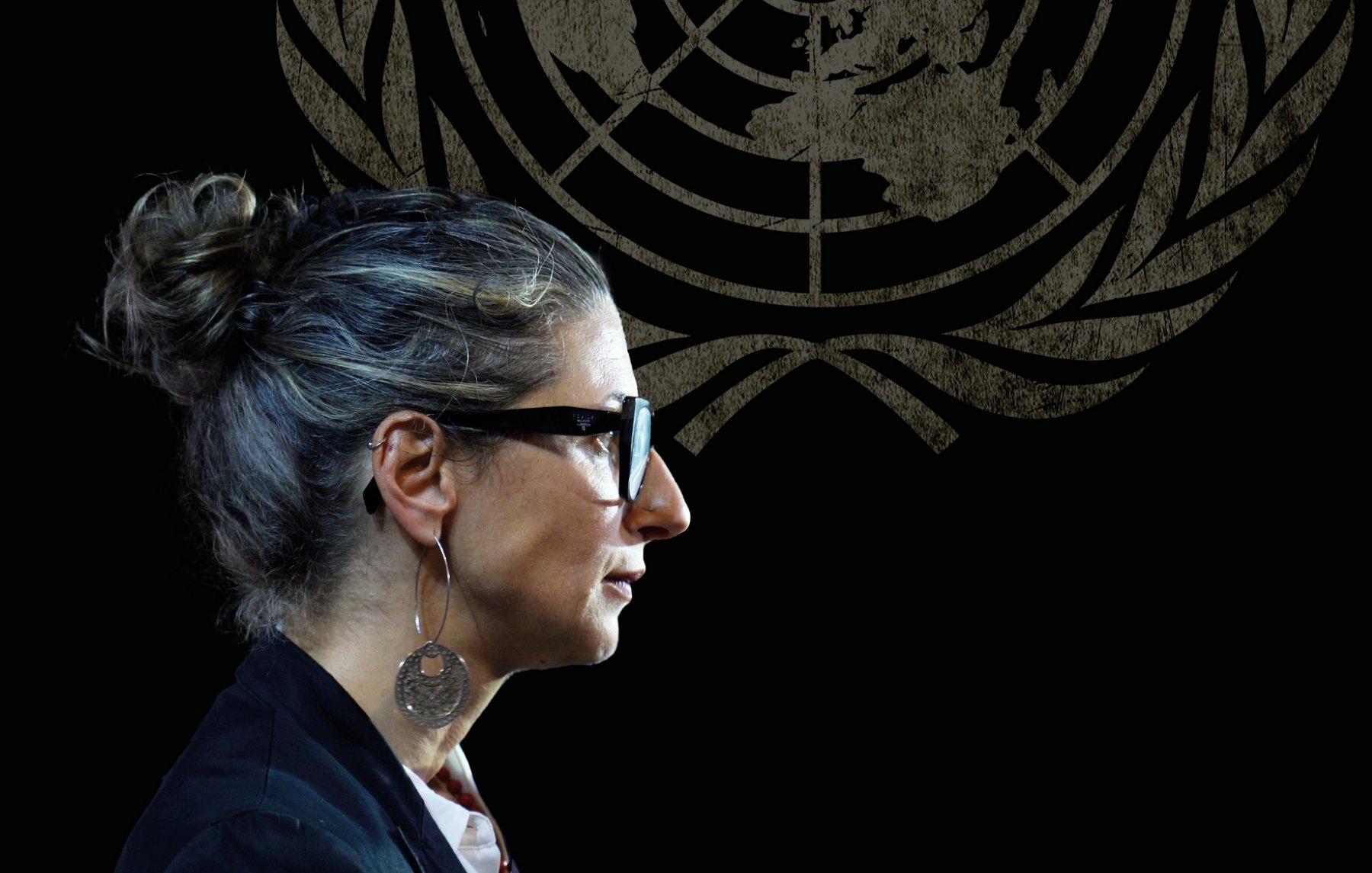 Donna con occhiali e capelli raccolti, in un ambiente con il logo delle Nazioni Unite sullo sfondo.