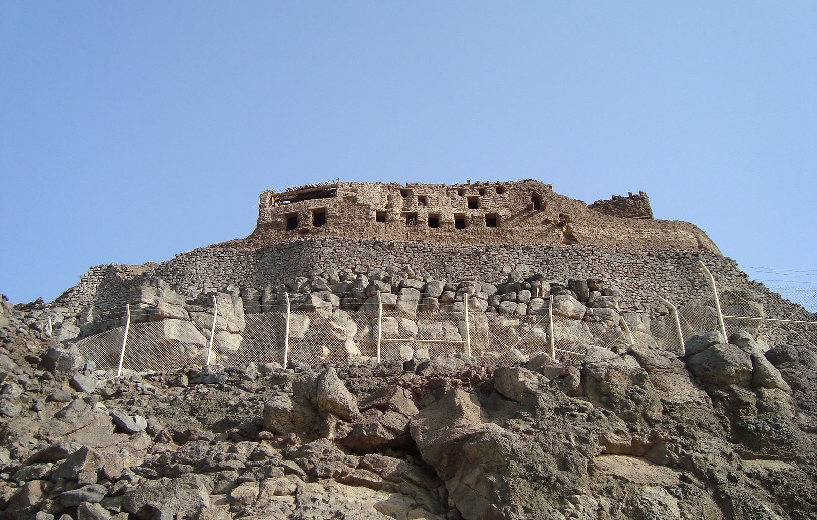 L'image montre une ancienne structure en pierre située sur une colline rocheuse. On peut voir des ruines dissimulées par des murs et des terrasses. Le ciel est dégagé, ce qui donne une belle luminosité à la scène. La végétation est absente, et le paysage est principalement composé de roches. La construction semble avoir une importance historique, suggérant qu'elle pourrait être un vestige d'une civilisation passée.