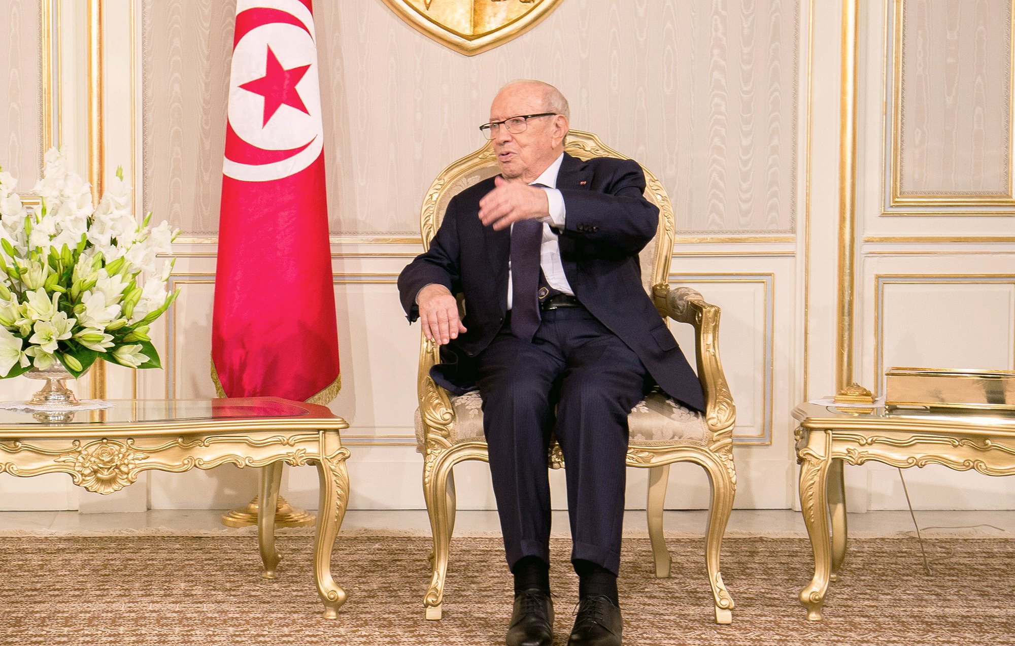 L'image montre un homme assis sur une chaise en or dans un environnement intérieur élégant. Il porte un costume sombre et des lunettes. À sa gauche, il y a un vase avec des fleurs blanches. Derrière lui, on aperçoit le drapeau tunisien. L'homme semble être en train de parler ou de gesticuler, indiquant une conversation ou une discussion en cours. Les murs et le décor environnant évoquent une atmosphère formelle et institutionnelle.