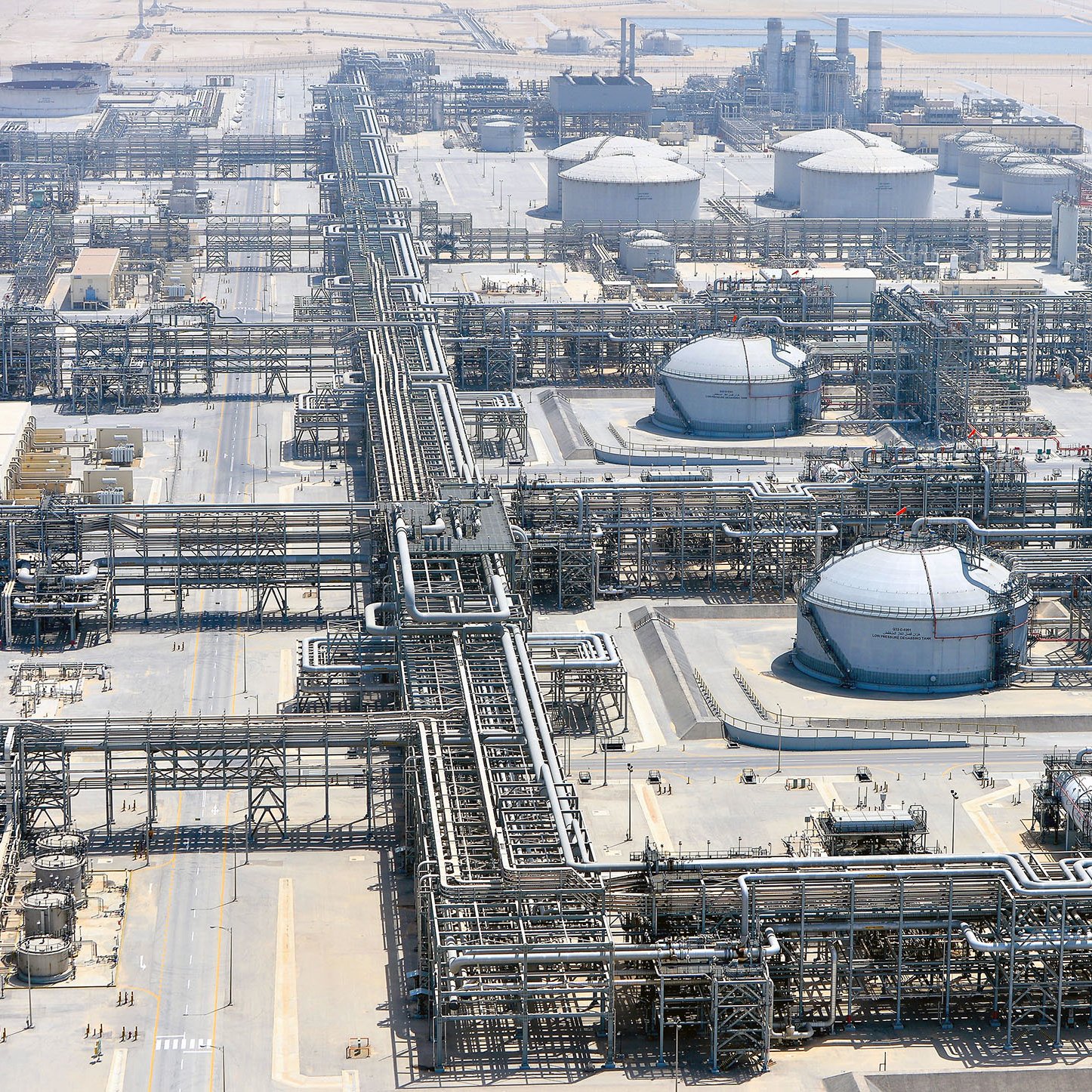 L'image montre une vue aérienne d'une installation industrielle, vraisemblablement une raffinerie ou un complexe pétrolier. On peut y voir des réservoirs de stockage de forme sphérique, entourés de nombreux tuyaux et structures métalliques. L'environnement est apparemment désertique, avec des terrains arides et peu de végétation. Plusieurs installations industrielles sont visibles, indiquant une activité de traitement ou de transport de matières pétrolières.