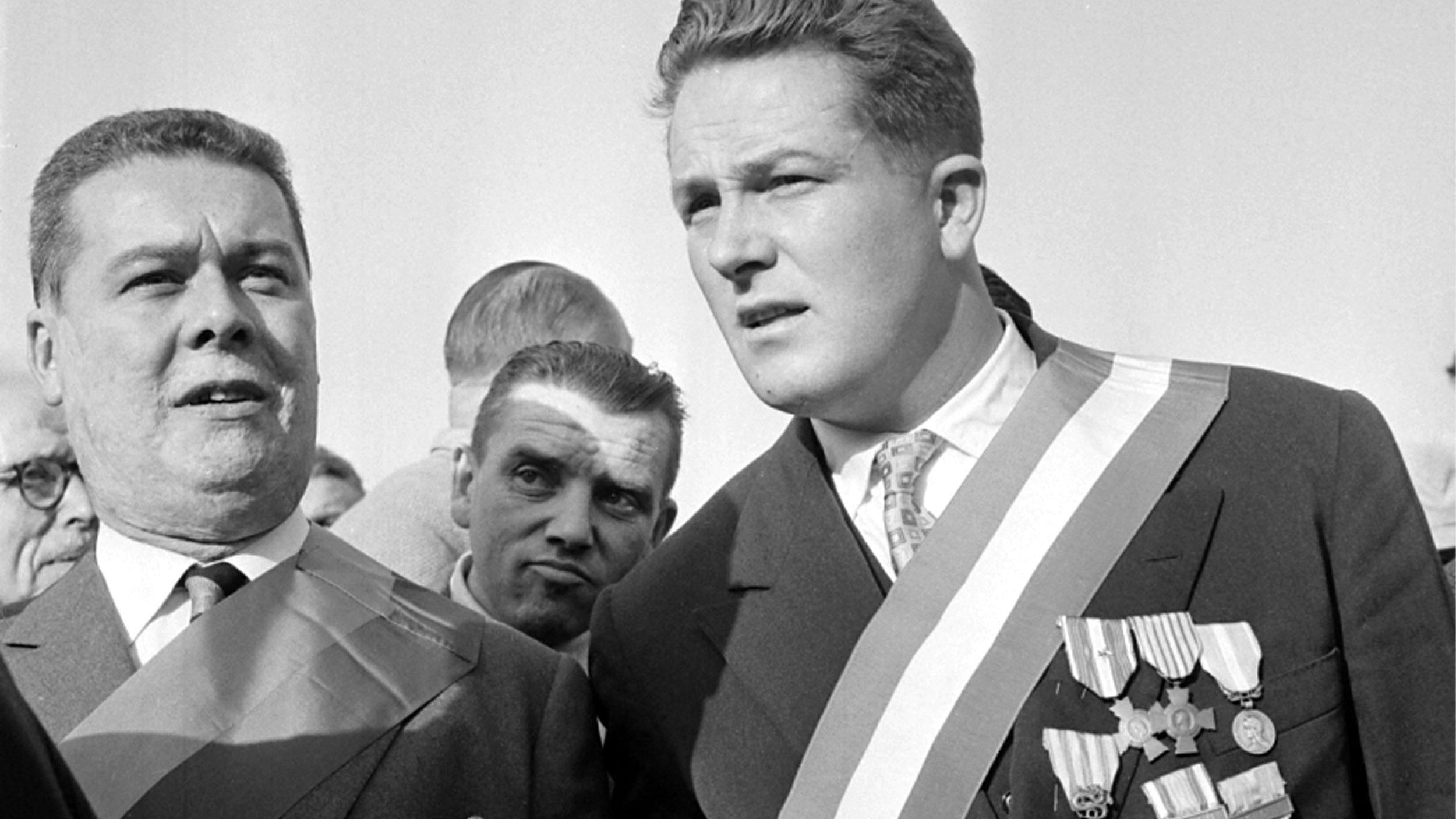 در این تصویر، دو مرد با لباس‌های رسمی و مدال‌های زیاد در حال صحبت یا مشاهده‌ی چیزی هستند. یکی از آن‌ها دارای مدال‌های متعددی بر روی سینه‌اش است و بر روی شانه‌اش نیز روبان بزرگی وجود دارد. مرد دیگر نیز به نظر می‌رسد مقام رسمی‌تری دارد و با دقت به طرف مقابل نگاه می‌کند. زمینه تصویر شامل افرادی دیگر است که به آنها توجه نمی‌شود. تصویر به وضوح فضایی رسمی و مربوط به یک مراسم یا رویداد مهم را نشان می‌دهد.