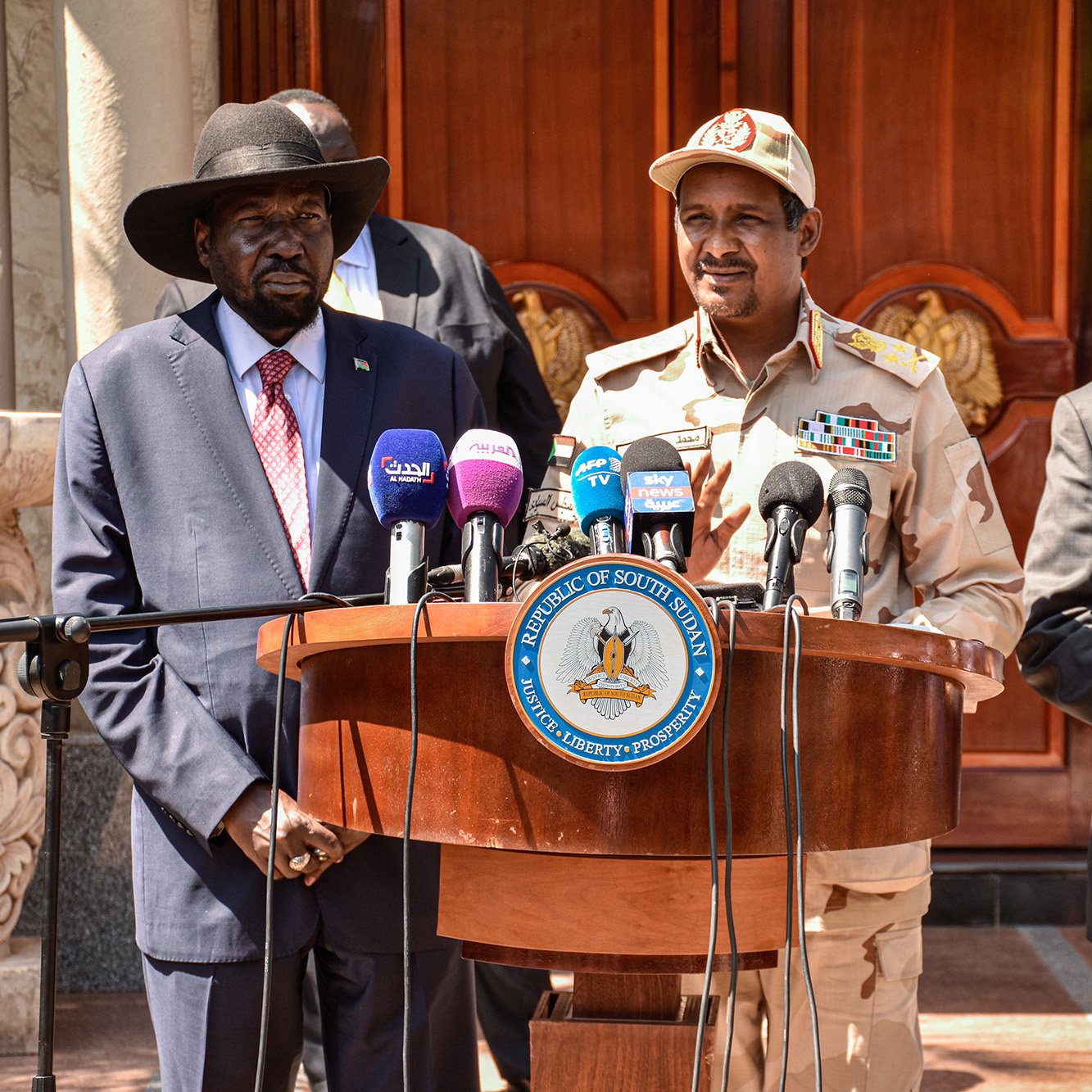 تظهر الصورة ثلاثة رجال يقفون أمام منصة. الشخص الموجود على اليسار يرتدي بدلة رسمية وقبعة عريضة، بينما الشخص في الوسط يرتدي زي عسكري وقبعة، والشخص في اليمين يرتدي بدلة رسمية أيضاً. يوجد خلفهم مبنى ذو تصميم معمارى مميز، ويبدو أنهم في مناسبة رسمية أو مؤتمر صحفي. المنصة تحتوي على شعارات وأعلام تُشير إلى الحكومة أو الدولة المعنية.