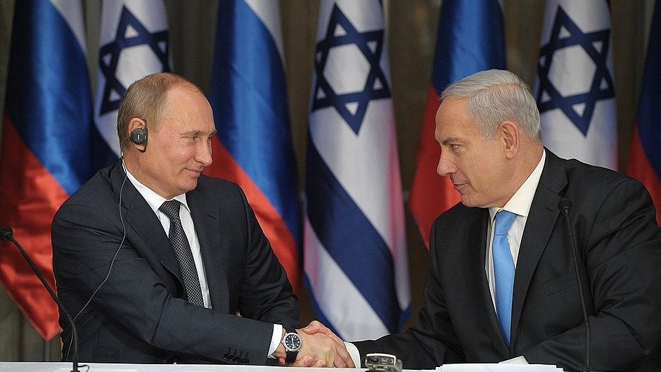 L'image montre deux hommes en costume, se serrant la main, dans un cadre formel. À leur arrière-plan, on peut voir des drapeaux de la Russie et d'Israël. L'un des hommes, à gauche, semble être en train de porter un écouteur, tandis que l'autre, à droite, regarde attentivement. L'ambiance suggère un moment de coopération ou d'accord.