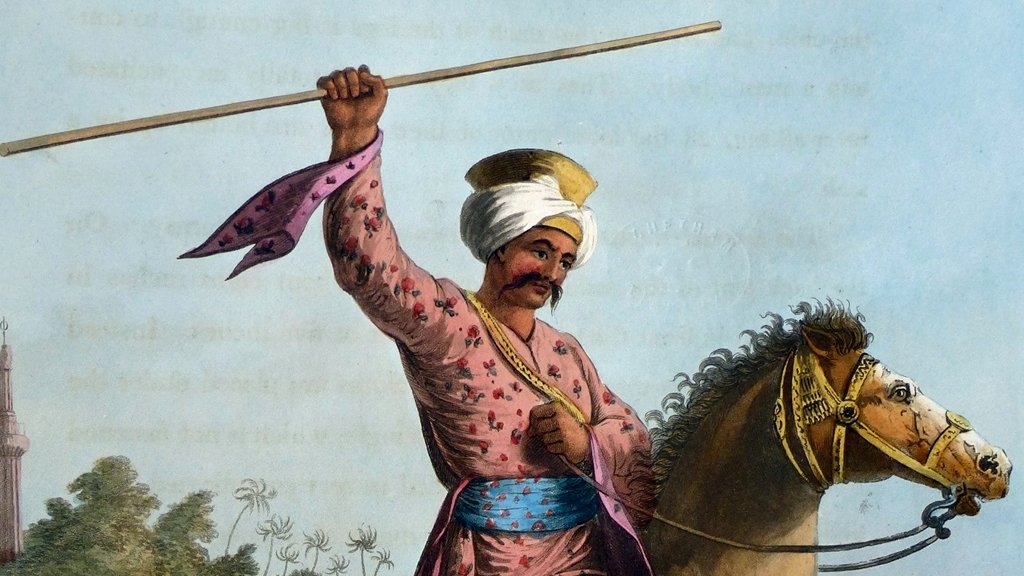 L'image représente un cavalier en tenue traditionnelle. Il porte un turban et des vêtements colorés ornés de motifs, avec une ceinture large. Le cavalier est en train de tenir un long bâton levé, semblant exprimer force et détermination. Il est monté sur un cheval puissant, qui avance au galop. En arrière-plan, on peut apercevoir d'autres cavaliers, ce qui suggère une scène dynamique. L'environnement semble indiquer un paysage désertique ou aride, renforçant le caractère historique de l'image.