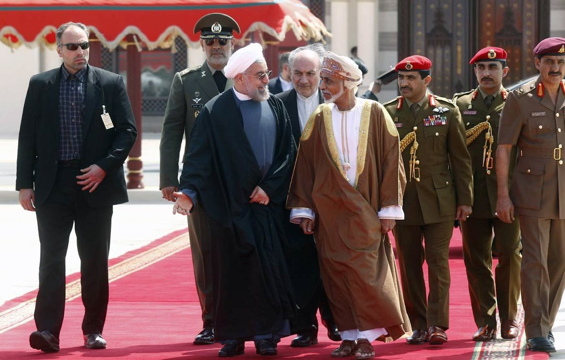 The image features a group of men walking together on a red carpet, likely in a formal or diplomatic setting. In the foreground, two men are engaged in conversation, one wearing a black suit and the other in traditional attire. They appear to be leaders or dignitaries. Behind them, there are several men in military uniforms and suits, possibly security personnel or officials, accompanying the two main figures. The background suggests an ornate architectural setting, enhancing the official nature of the gathering.