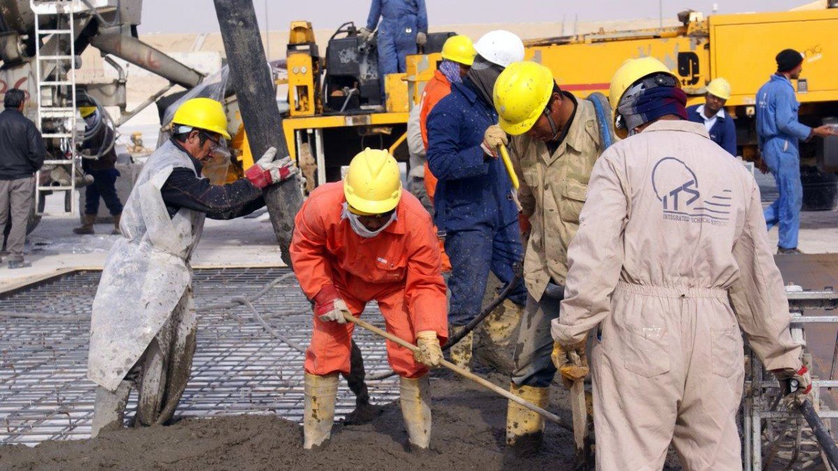در این تصویر گروهی از کارگران مشغول به کار در یک پروژه ساختمانی هستند. آنها کلاه ایمنی به سر دارند و لباس‌های کار پوشیده‌اند. چند نفر در حال ریختن بتن و استفاده از تجهیزات مختلف هستند. محیط کار شلوغ و صنعتی به نظر می‌رسد و ماشین‌آلاتی در پس‌زمینه دیده می‌شود. کارگران با جدیت و همکاری در حال انجام وظایف خود هستند.