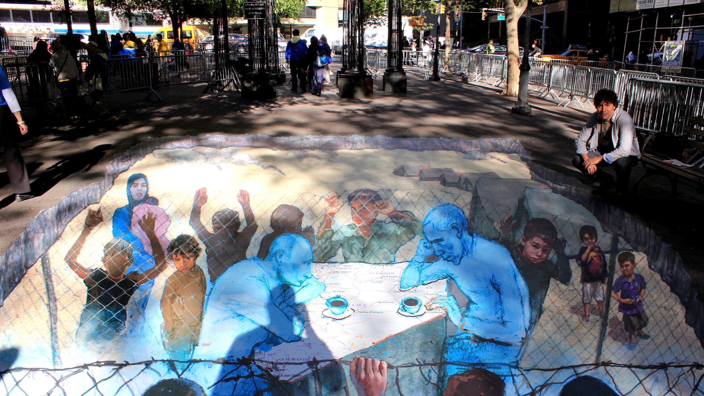 L'image montre une œuvre d'art en 3D peinte sur le sol, représentant des figures humaines engagées dans une conversation. On y aperçoit deux hommes assis à une table, entourés de scènes de détresse, symbolisant possiblement des personnes en situation de crise. Des enfants et d'autres figures semblent observer, créant un fort impact émotionnel. La couleur bleue prédomine, ajoutant à la gravité de la scène. L'environnement urbain autour de l'œuvre indique que cela se déroule dans un espace public, où des passants peuvent réfléchir sur le message véhiculé par cette peinture.