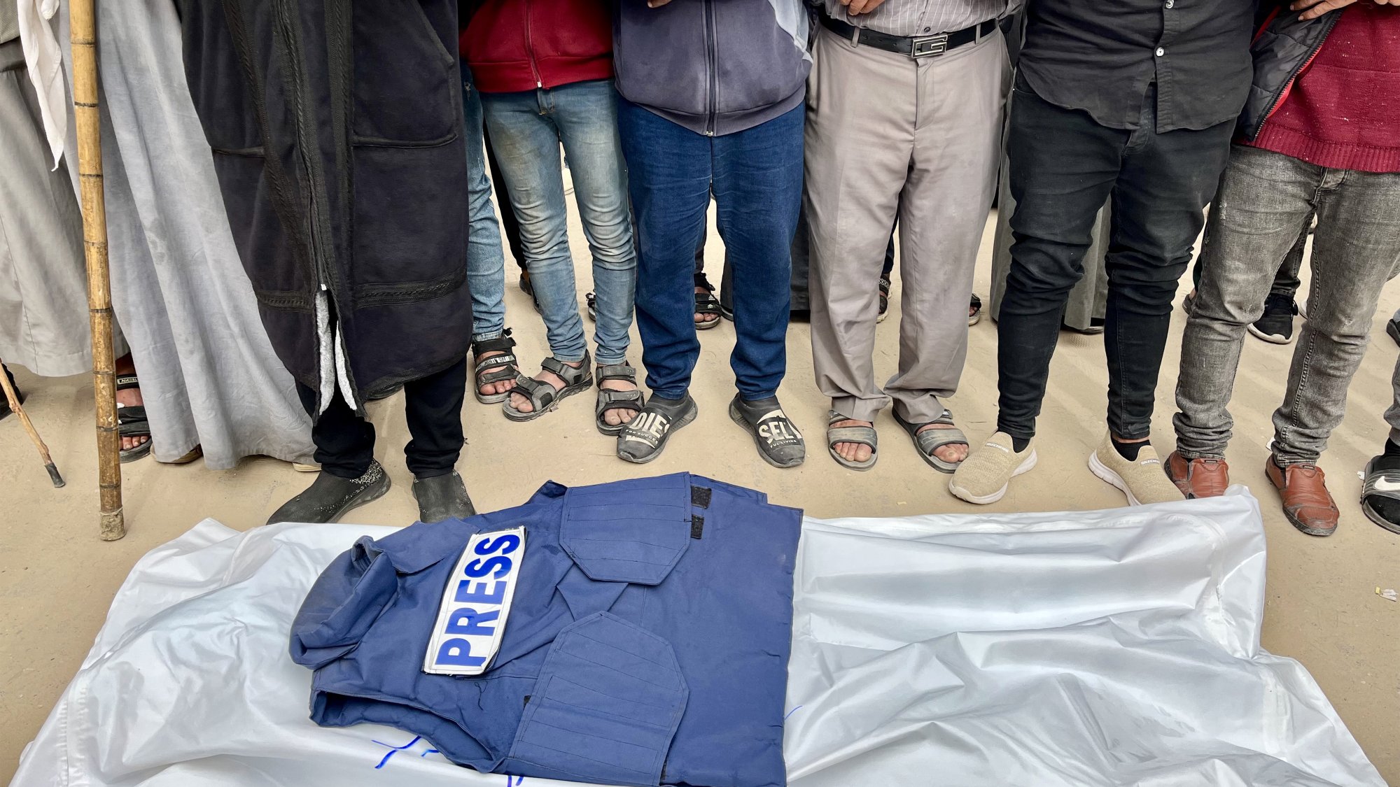 Des hommes entourent un gilet "PRESS" sur une bâche, en posture de recueillement.