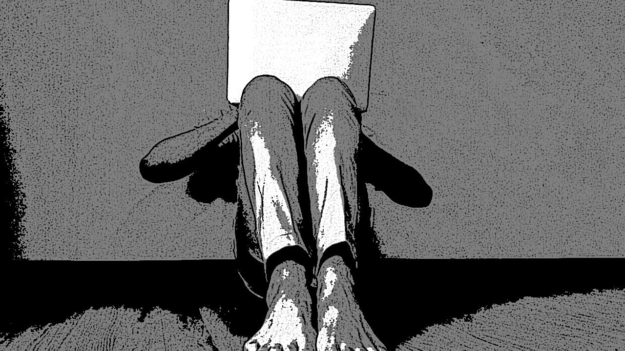 L'image montre une personne assise par terre, adossée à un mur. Elle tient un ordinateur portable devant elle, ce qui cache son visage. Ses jambes sont repliées et ses pieds nus sont visibles. L'ambiance est plutôt sombre et minimaliste, avec un contraste marqué entre les ombres et la lumière.