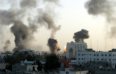L'image montre une vue d'une ville en ruines, avec plusieurs colonnes de fumée s'élevant dans le ciel au crépuscule. Les bâtiments sont partiellement détruits, et l'atmosphère semble tendue et chaotique. La lumière du coucher de soleil se mêle aux nuages de fumée, créant un contraste marqué entre la lumière et l'obscurité. Cela évoque une scène d'urgence ou de conflit.