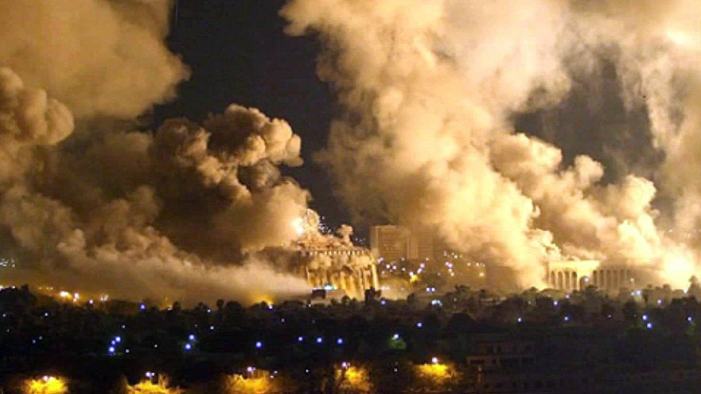 L'image montre une scène nocturne dramatique avec des nuages de fumée épais s'élevant vers le ciel. On peut voir des structures lumineuses en arrière-plan, suggérant que des bâtiments sont présents, mais ils sont partiellement obscurcis par la fumée. La réflexion des lumières sur une surface d'eau au premier plan ajoute une atmosphère sombre et intense à la composition. L'ensemble évoque une situation de conflit ou de désastre.