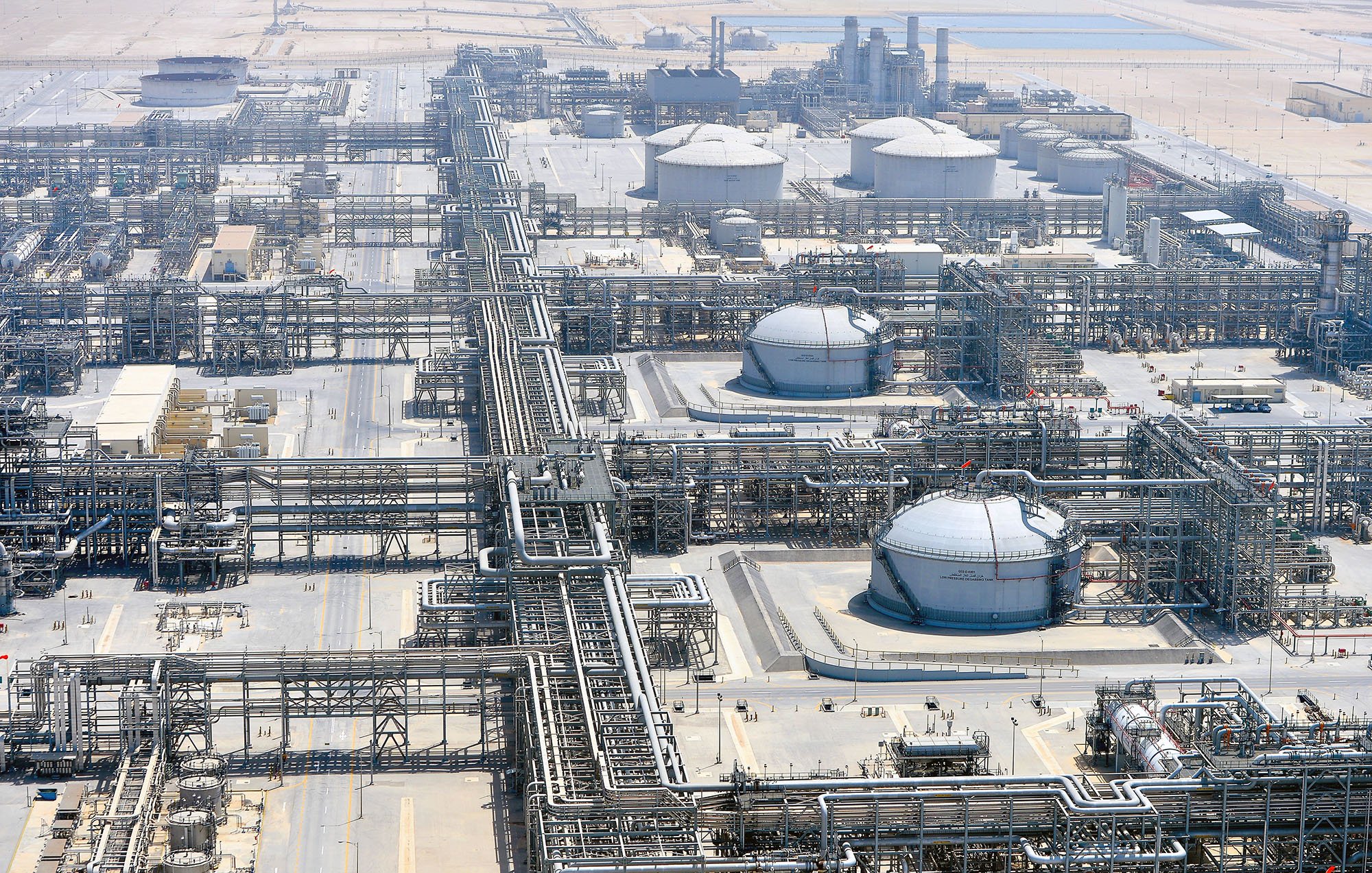 The image depicts a large industrial facility, likely an oil refinery or petrochemical plant. It features numerous large storage tanks, interconnected pipelines, and extensive infrastructure. The site is expansive, suggesting significant industrial activity, and it appears to be located in a relatively arid region, as indicated by the surrounding landscape. There are various structures for processing and storage, emphasizing the complexity and scale of the operation.
