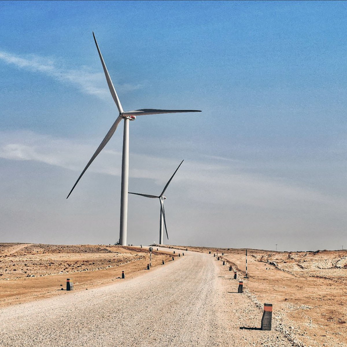 تظهر في هذه الصورة منظرًا طبيعيًا صحراويًا يمتد تحت سماء زرقاء فاتحة. يتواجد في الجزء الأمامي طريق رملي يتجه نحو الأفق، محاطًا بأعمدة صغيرة على جانبيه. في الخلفية، هناك عدة توربينات رياح ضخمة تدور ببطء، مما يُعطي انطباعًا حديثًا حول استخدام الطاقة المتجددة في هذا المناطق القاحلة. يبدو المكان هادئًا وواسعًا، مع تباين واضح بين الألوان الرمادية للأرض والألوان الزرقاء للسماء.