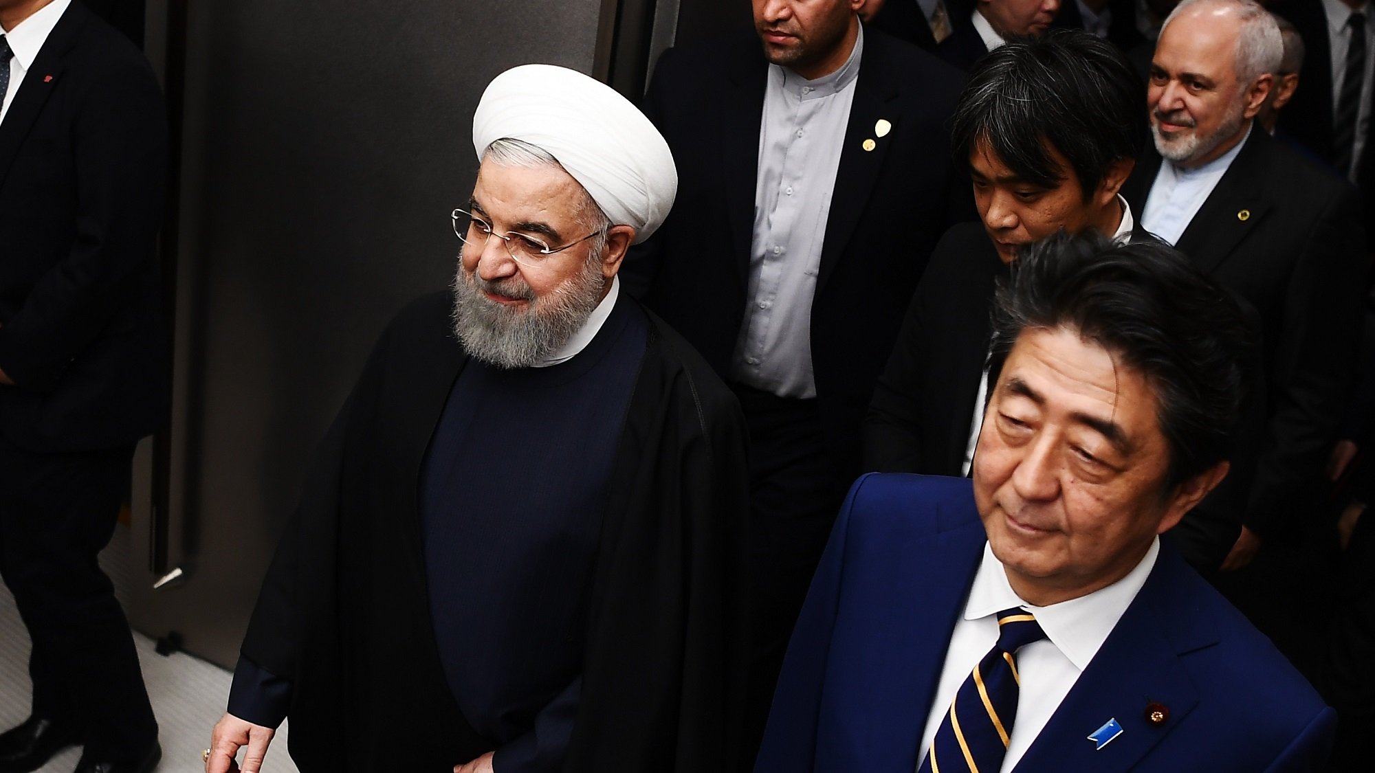 در تصویر، دو مرد در حال پیاده‌روی هستند. یکی از آنها دارای پوشش سنتی ایرانی با عمامه سفید و جبه است، و به نظر می‌رسد که او مقام رسمی و بلندپایه‌ای باشد. فرد دیگر در کنار او، با کت و شلوار رسمی و کراوات، لبخند به لب دارد. در پس‌زمینه، چند نفر دیگر نیز دیده می‌شوند که به نظر می‌رسد همراهان آنها هستند. محیط تصویر ممکن است یک کنفرانس یا جلسه رسمی را نشان دهد.