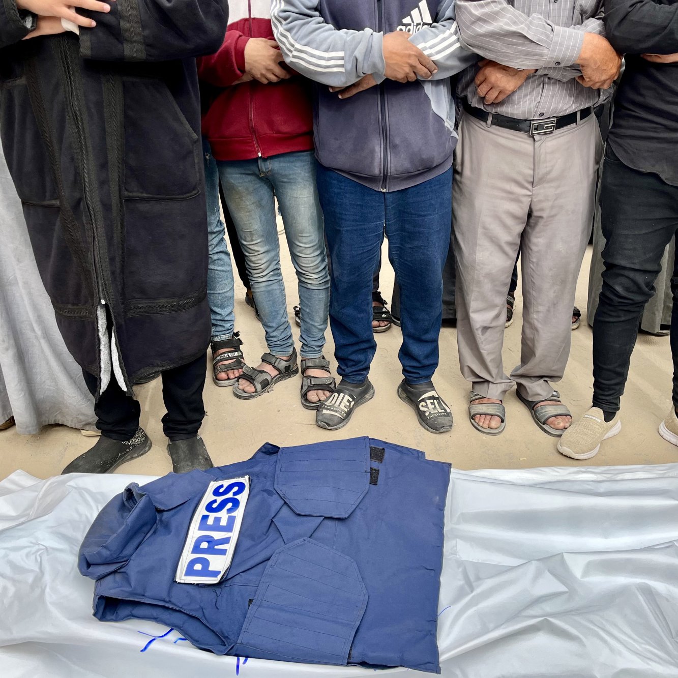 Des hommes entourent un gilet "PRESS" sur une bâche, en posture de recueillement.