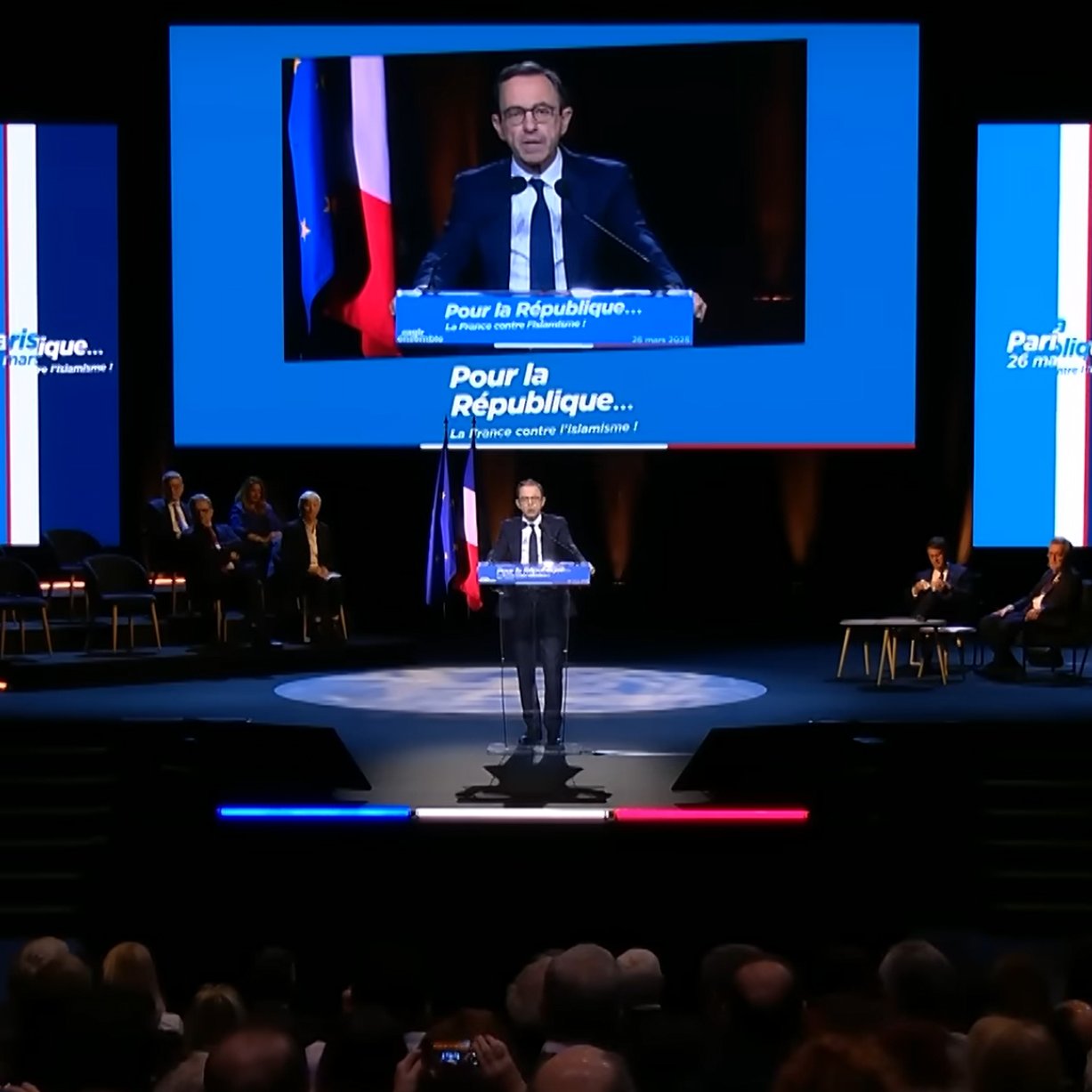 Un homme parle sur scène, avec des drapeaux français en arrière-plan. 