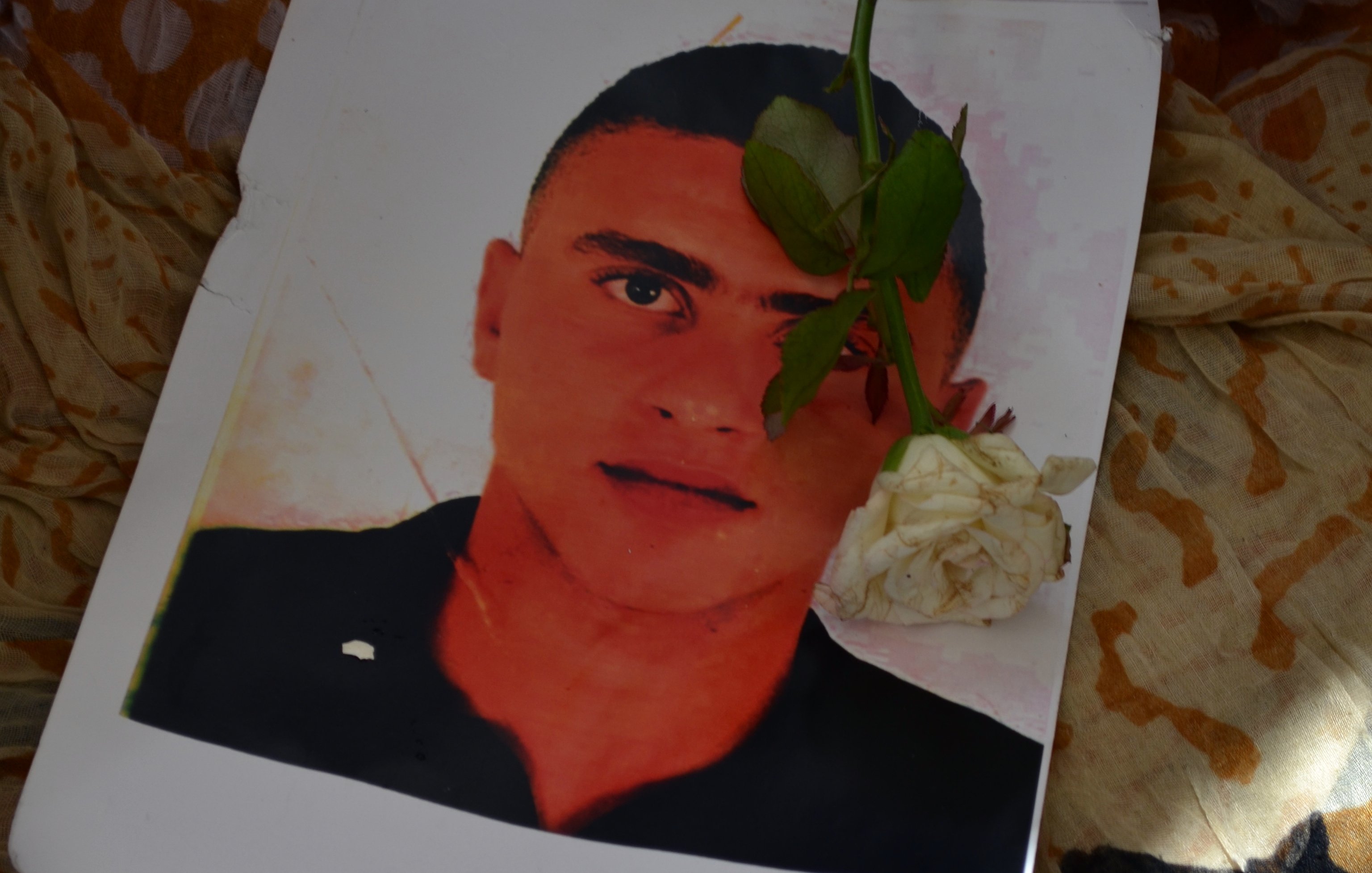 تظهر في الصورة ورقة مطبوعة تحتوي على صورة شاب، حيث يوضع عليها وردة بيضاء. الخلفية تبدو ملونة ومعقدة، مما يضيف جوًا من الحزن والحنين. الورقة قد تشير إلى ذكرى لهذا الشخص، والزهرة تعزز من مشاعر التأمل والاحترام.