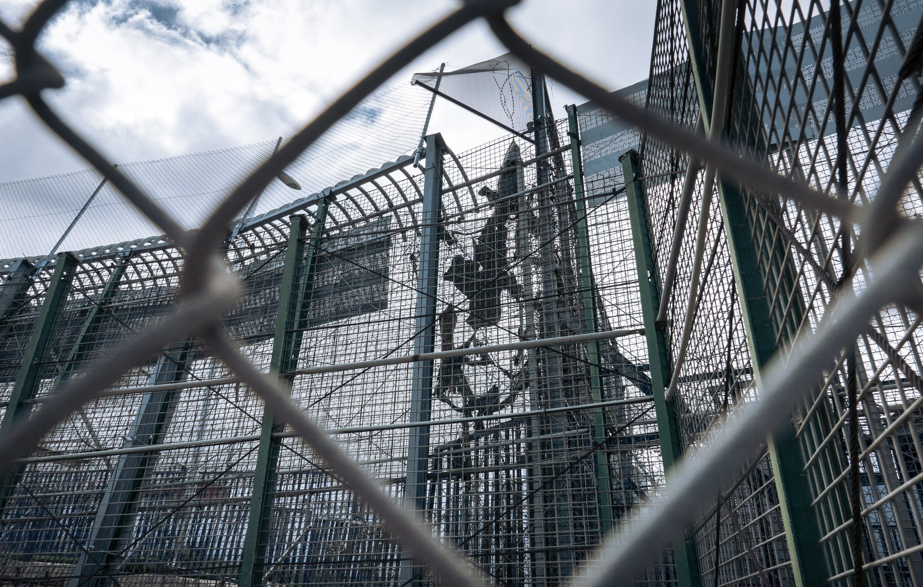 L'image montre une structure en métal, probablement une clôture ou un mur de sécurité, entourée de grillages. On peut apercevoir des barres et une superficie qui semble être une installation sécurisée. Le ciel est nuageux, ce qui donne une atmosphère un peu sombre à la scène. Cette image évoque un environnement restreint, probablement lié à la sécurité ou à la protection.