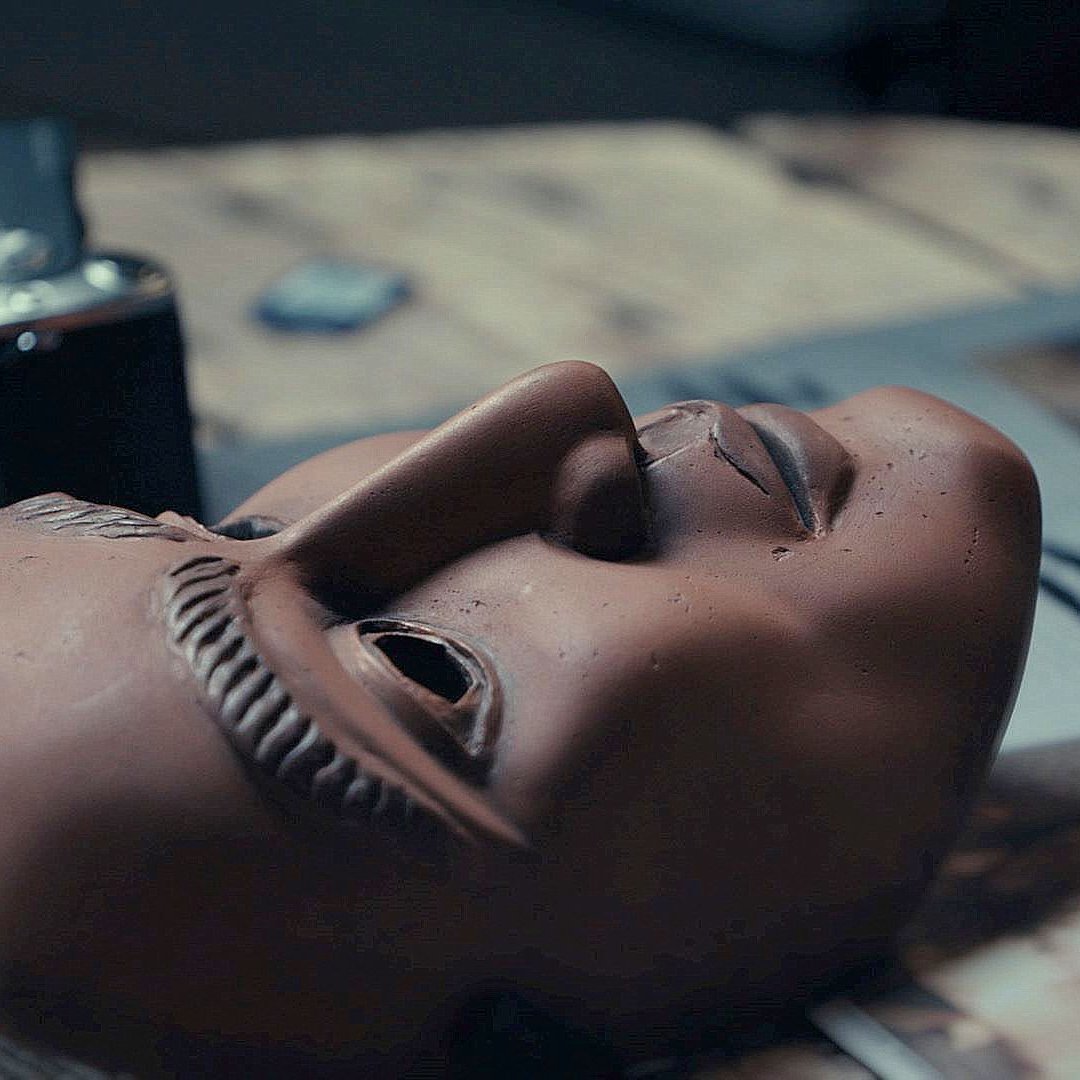 L'image montre un masque en terre cuite posé sur une surface en bois. Le masque a des traits détaillés, avec une expression neutre et des sourcils sculptés. À côté du masque, on voit une montre ou un objet similaire, ainsi qu'un magazine ouvert avec des illustrations. L'éclairage est doux, créant une ambiance contemplative autour de ces objets.