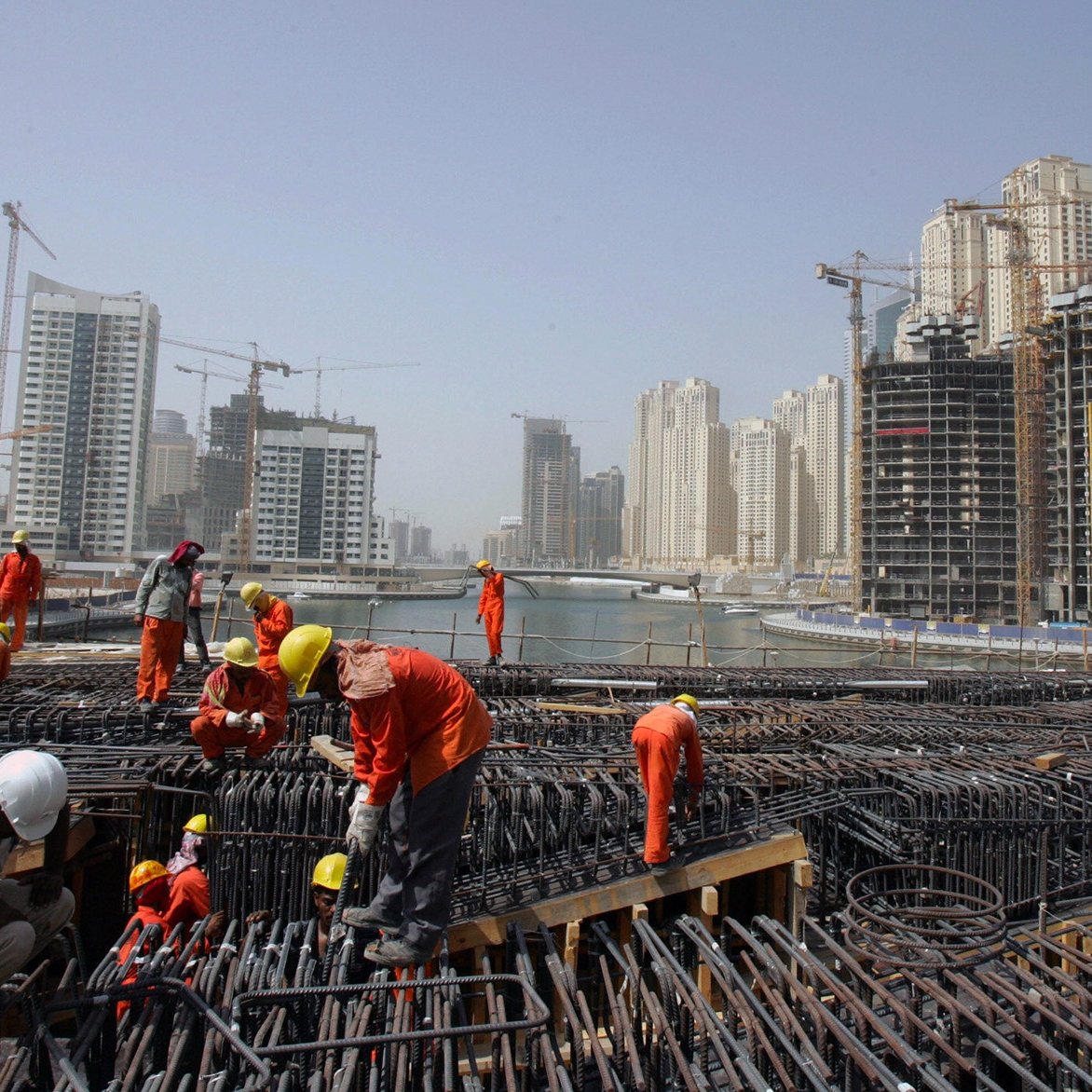L'image montre un chantier de construction animé, où de nombreux ouvriers en uniformes orange et casques de sécurité s'affairent autour de structures en acier. En arrière-plan, on peut voir des immeubles modernes et des grues, illustrant une zone urbaine en développement. Le ciel est clair, et l'ensemble dégage une atmosphère dynamique de construction.