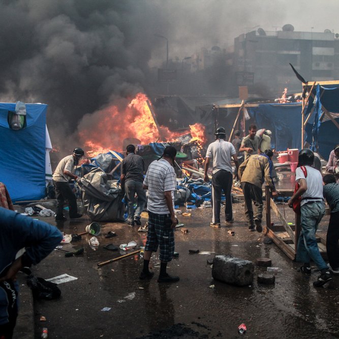 L'image montre une scène de chaos et de destruction. On peut voir des personnes rassemblées autour de flammes, visiblement en train de fuir ou de lutter contre un incendie. La fumée noire s'élève dans le ciel, obscurcissant la vue. Des tentes et des objets sont dispersés dans l'environnement, suggérant une occupation ou une manifestation. La situation semble tendue, avec un sentiment d'urgence parmi les personnes présentes.