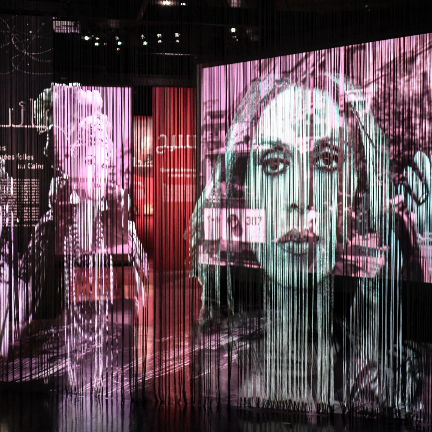 L'immagine presenta una serie di proiezioni artistiche su uno sfondo di tende di tessuto trasparente. Le immagini ritraggono volti femminili e scene di vita, con un forte contrasto tra bianco e nero e tonalità colorate. L'atmosfera è suggestiva e intimista, creando un effetto di profondità e movimento. Le fotografie sembrano evocare un senso di nostalgia, magari legato alla storia e alla cultura. Le scritte in arabo e altre lingue suggeriscono un tema culturale e un messaggio profondo.