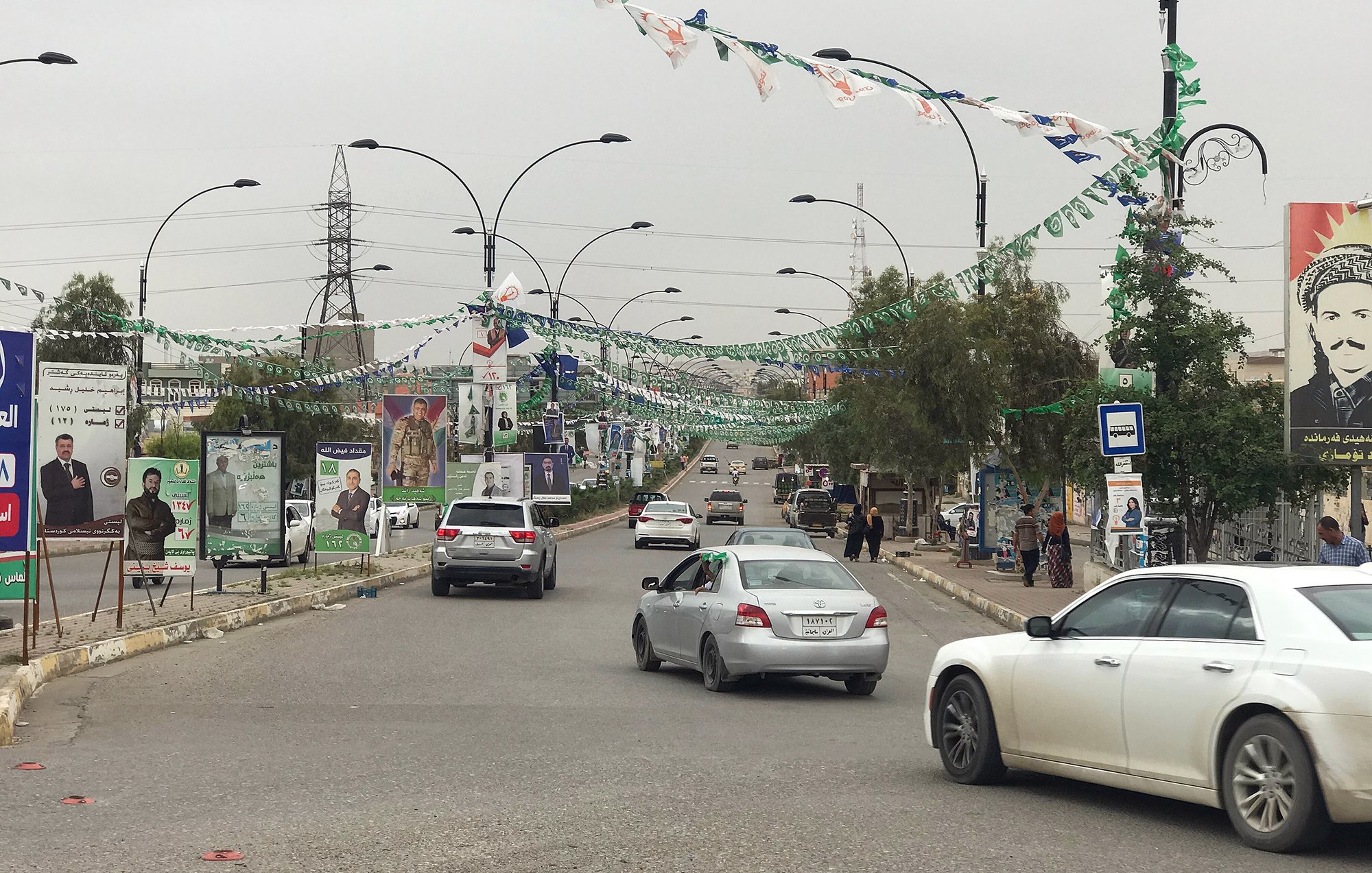 L'image montre une rue bordée de drapeaux et de bannières, probablement en préparation d'un événement ou d'une célébration. On peut voir plusieurs voitures circulant sur la route. À côté de la route, il y a des panneaux avec des affiches, ce qui indique une zone urbaine active. Des personnes se trouvent également sur le trottoir, et des arbres et des lampadaires sont présents le long de la rue, donnant un aspect dynamique à la scène. Le ciel semble nuageux, suggérant une ambiance calme.