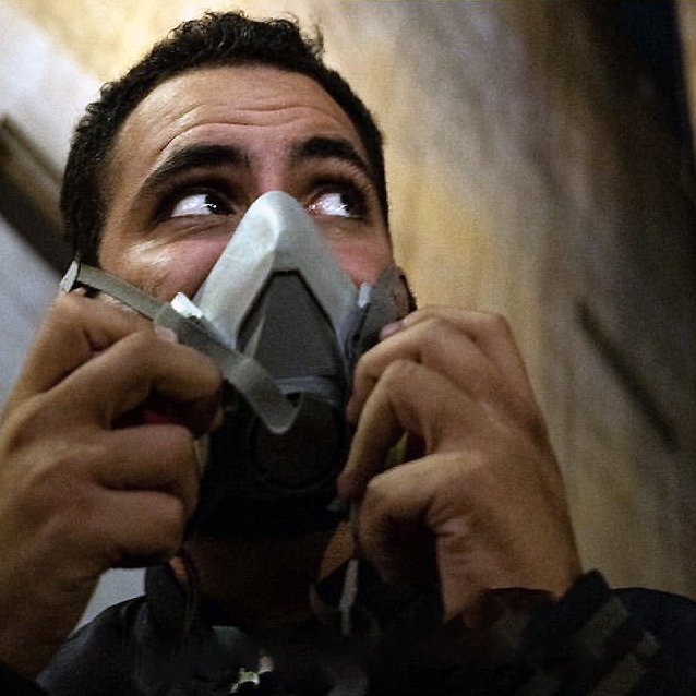 L'image montre un homme qui porte un masque à gaz. Son visage exprime une certaine vigilance, et il semble regarder autour de lui. L'environnement est urbain, avec des murs et des bâtiments en arrière-plan. L'éclairage donne une ambiance sombre et intense à la scène.