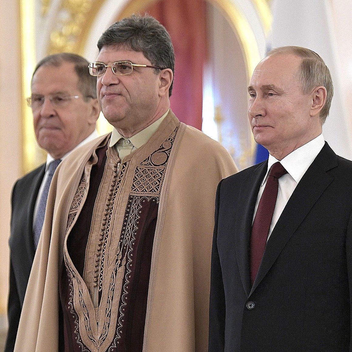 L'image montre trois hommes en pleine cérémonie. Au centre, un homme porte une tenue traditionnelle ornée de motifs. À sa gauche, un homme en costume formel et à sa droite, un homme aussi en costume. Le décor semble somptueux avec des dorures et des drapeaux en arrière-plan, suggérant un lieu officiel. L'atmosphère semble sérieuse et formelle.