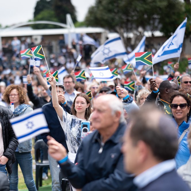 در این تصویر، گروهی از مردم در یک مراسم یا تجمع بزرگ دیده می‌شوند. آن‌ها در حال تکان دادن پرچم‌های اسرائیل و آفریقای جنوبی هستند. جمعیت شامل مردان و زنانی از تنوع سنی مختلف است و برخی از آن‌ها در حال لبخند زدن و خوشحالی دیده می‌شوند. فضای عمومی کمی ابری به نظر می‌رسد و تعداد زیادی کرسی در پس‌زمینه وجود دارد.
