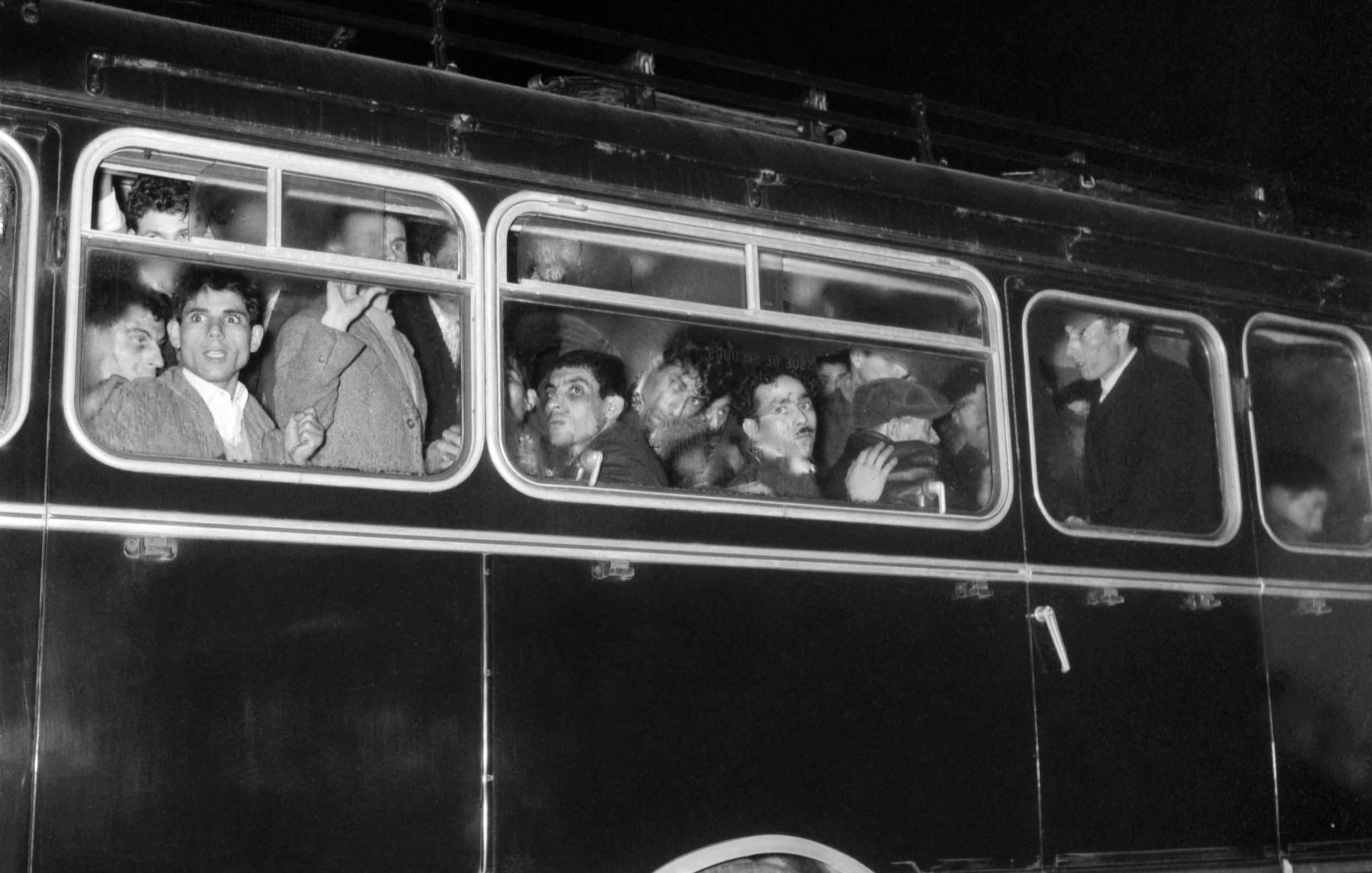 تظهر الصورة حافلة قديمة في الليل، وهي مزدحمة بالركاب الذين يبدون في حالة من الحماس أو الانفعالات. بعض الأشخاص ينظرون من النوافذ في اتجاه الكاميرا، بينما يبدو الآخرون مشغولين بالحديث أو التفاعل مع بعضهم. الأضواء الخافتة تضفي جواً مميزاً على المشهد، مما يعكس لحظة مميزة أو حدثاً معيناً. الحافلة تتميز بتصميمها الكلاسيكي، مما يشير إلى فترة زمنية سابقة.