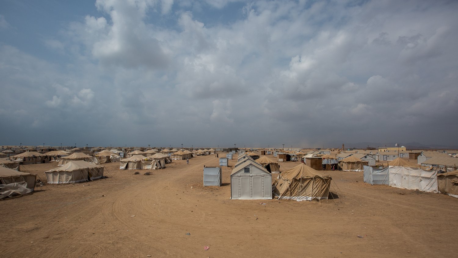 تظهر في الصورة مشهدًا صحراويًا واسعًا، حيث تنتشر العديد من الخيام على الأرض الرملية. السماء ملبدة بالسحب، مما يوحي بوجود طقس متغير. يبدو أن المكان يحمل طابعًا يجمع بين البساطة والاحتياجات الأساسية، حيث تُستخدم الخيام كوسيلة للإيواء. تبرز بعض الخيام بألوان فاتحة ومختلفة، مما يضفي طابعًا متنوعًا على المنطقة.