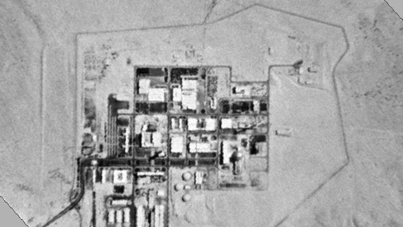 L'image montre une vue aérienne en noir et blanc d'une installation industrielle ou militaire située dans une zone désertique. On peut y voir plusieurs bâtiments et structures disposés de manière ordonnée, entourés d'une clôture ou d'une barrière. Le paysage environnant semble aride, avec peu de végétation visible. Cette configuration suggère un site ayant des activités techniques ou de recherche avancées.