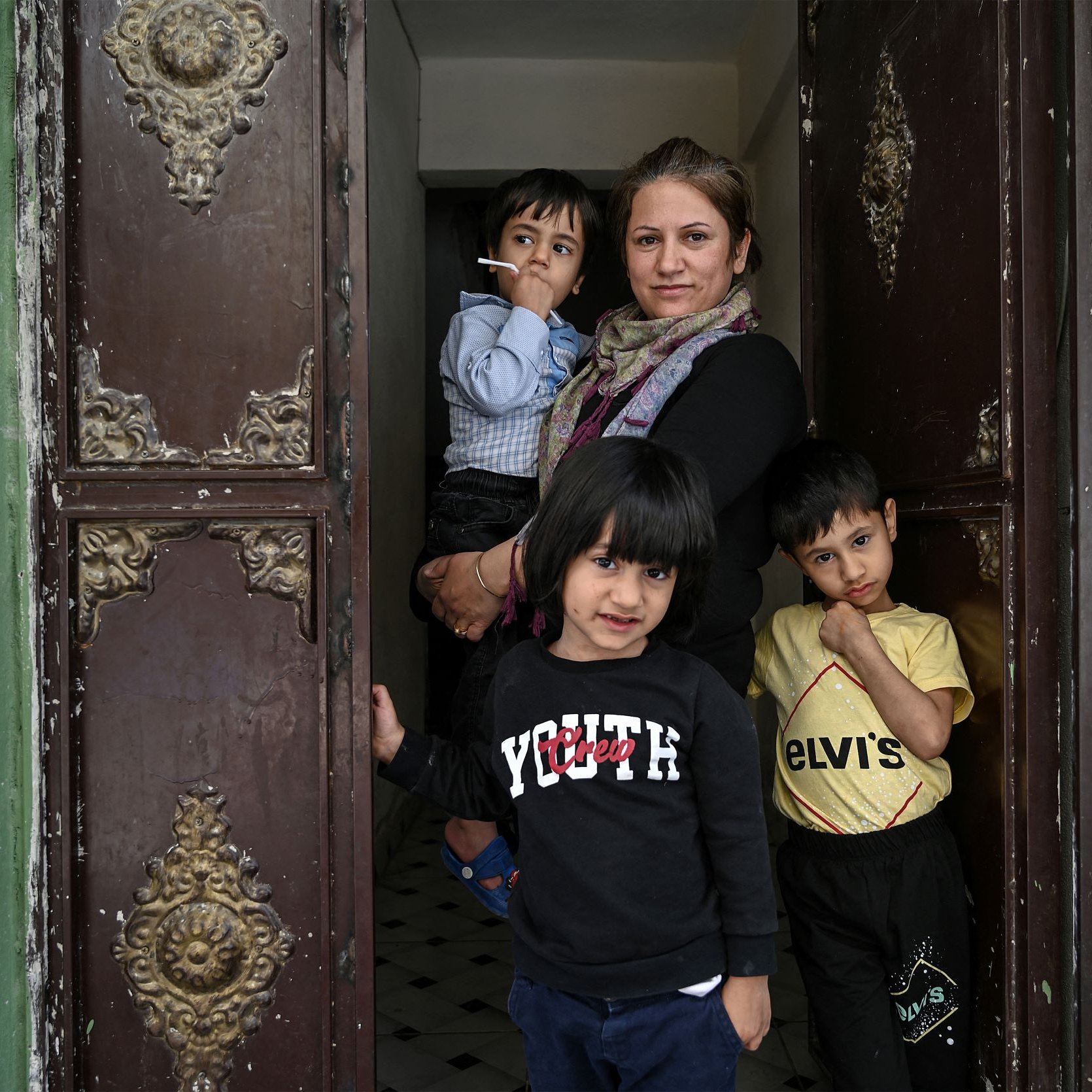 في الصورة، تظهر امرأة واقفة في مدخل منزل، تحمل سيجارة في يدها. خلفها، يوجد طفلان يلعبان، بينما يظهر طفل ثالث يقف أمامها. جدران المدخل ذات لون أخضر، والزخارف على الأبواب تظهر طابعًا قديمًا. تعكس تعابير الوجه في الصورة مزيجًا من الحذر والبراءة.