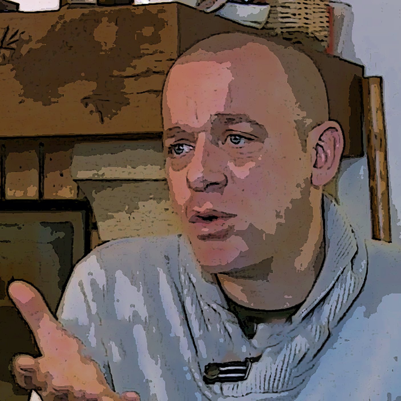 L'image montre un homme avec une coupe de cheveux courte et un pull en tricot. Il est assis près d'une cheminée en pierre. Son expression faciale semble être sérieuse, et il gesticule avec ses mains, ce qui suggère qu'il est en train d'expliquer ou de discuter d'un sujet important. L'arrière-plan présente des éléments décoratifs, ce qui ajoute une ambiance chaleureuse à la scène.