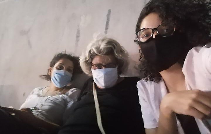 Sur l'image, on peut voir trois personnes assises côte à côte, chacune portant un masque. La première personne à gauche a les cheveux bouclés et porte un haut clair. La personne au milieu a des cheveux gris et porte des lunettes ainsi qu'un haut noir. La dernière personne à droite a des cheveux bouclés et porte également un haut clair. Elles semblent se détendre ensemble, peut-être dans un environnement public ou en extérieur. L'ambiance est informelle et conviviale.