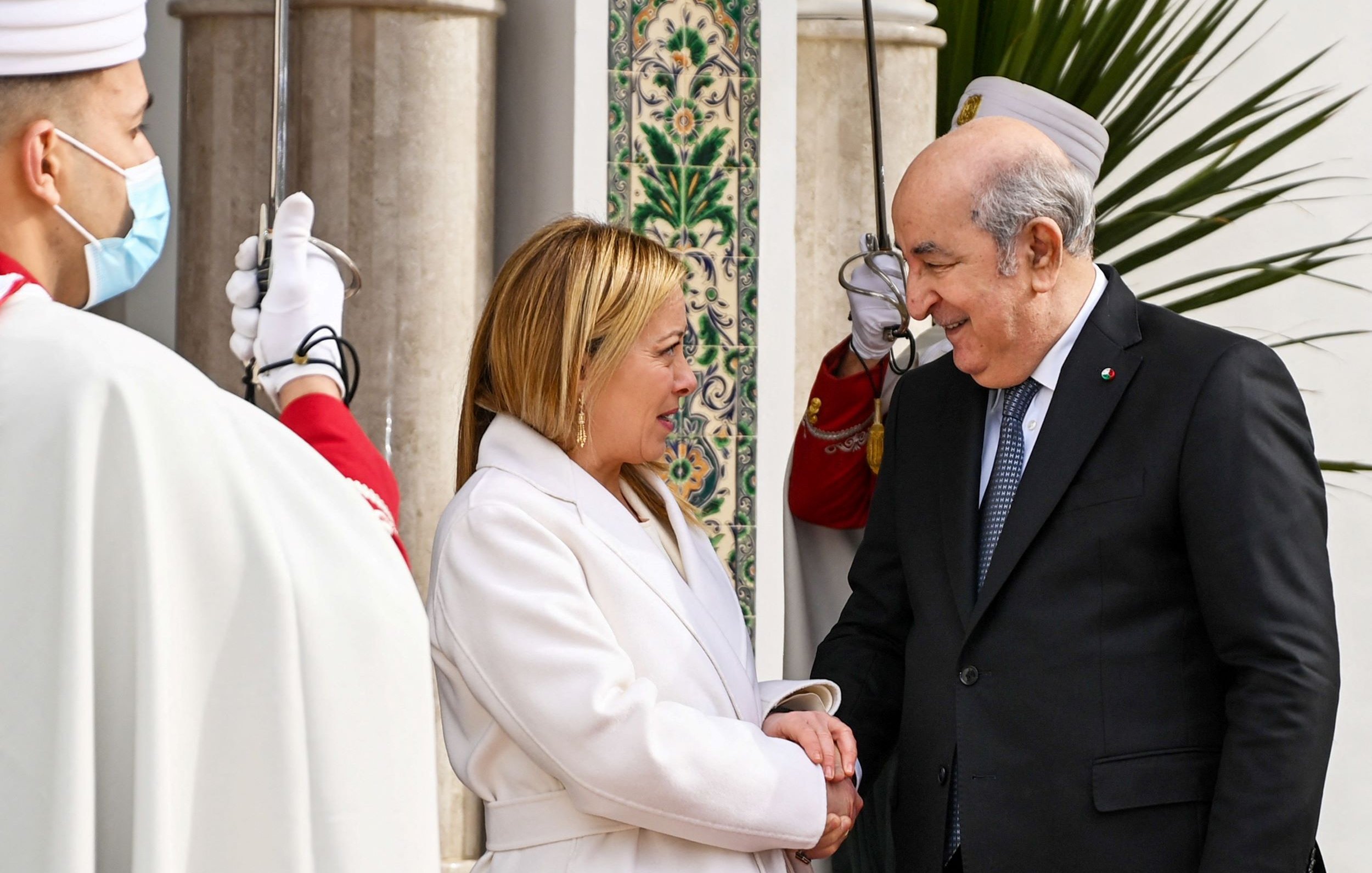 Nell'immagine si possono vedere due persone che si stringono la mano in un contesto formale. Una delle persone indossa un cappotto bianco e ha un sorriso amichevole, mentre l'altra è vestita in modo elegante, con un atteggiamento accogliente. Sullo sfondo si intravedono dettagli architettonici e persone in abiti tradizionali, creando un'atmosfera cerimoniale. La scena riflette un incontro ufficiale, suggerendo un momento di cordialità e dialogo.